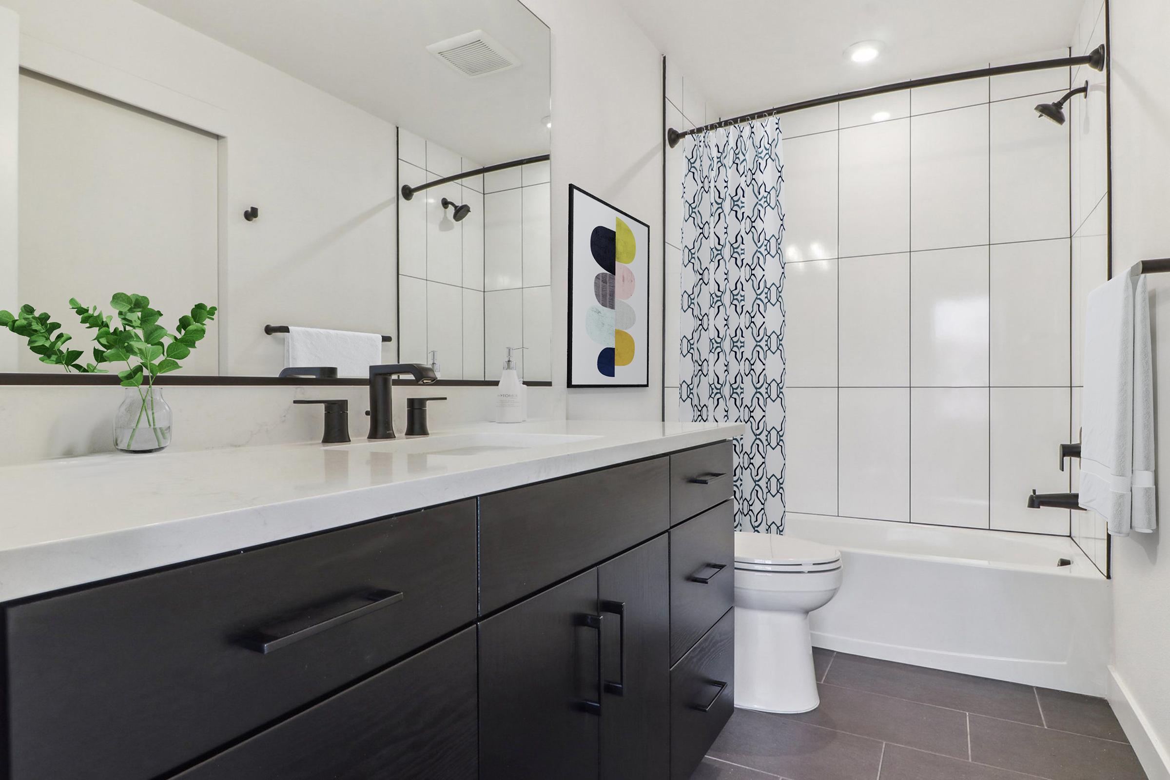 Modern bathroom featuring a black vanity with sleek drawers, a white countertop, and black faucets. A mirrored wall reflects the space, while a stylish shower curtain adds a pop of pattern. The bathtub is integrated into the design, and there are plants and decorative elements for a fresh look.