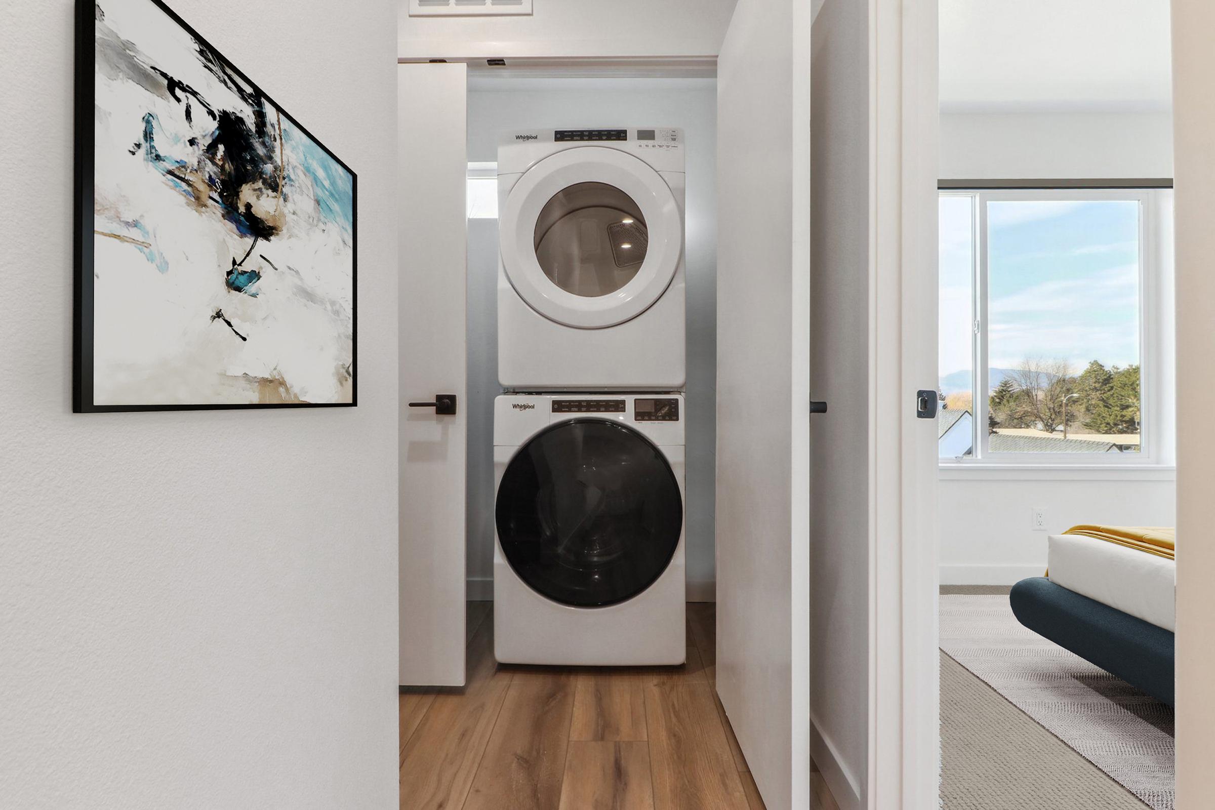 A modern laundry area featuring a stacked washer and dryer neatly positioned between two white walls, with a framed abstract painting on the left. Natural light filters in through a nearby window, highlighting the clean, contemporary design of the space.