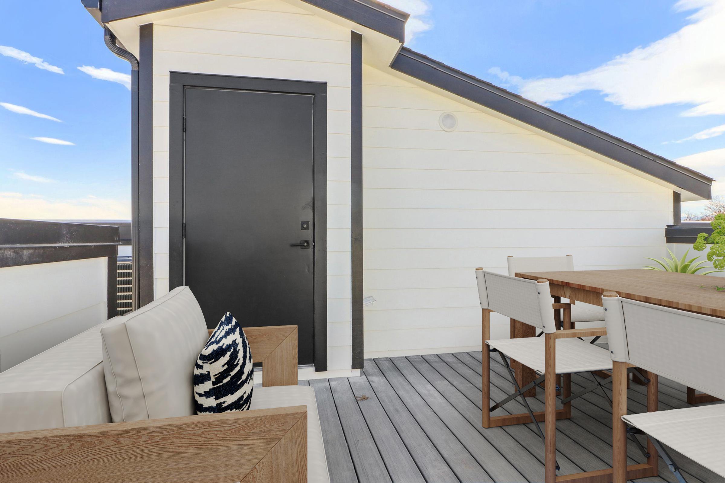 A modern outdoor patio featuring a minimalist design, with a black door, wooden table and chairs, and light-colored sofa cushions. The space is brightened by a blue sky and clouds, creating an inviting atmosphere for outdoor gatherings or relaxation.