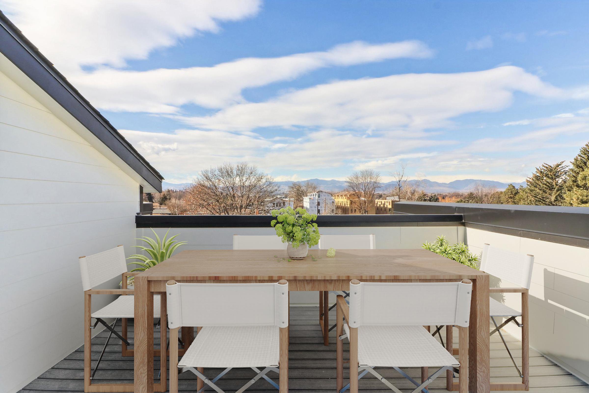 A spacious rooftop terrace featuring a wooden dining table surrounded by white chairs. The setting is enhanced by potted plants, with a panoramic view of trees and distant mountains under a clear blue sky with scattered clouds. Ideal for outdoor dining or relaxation.