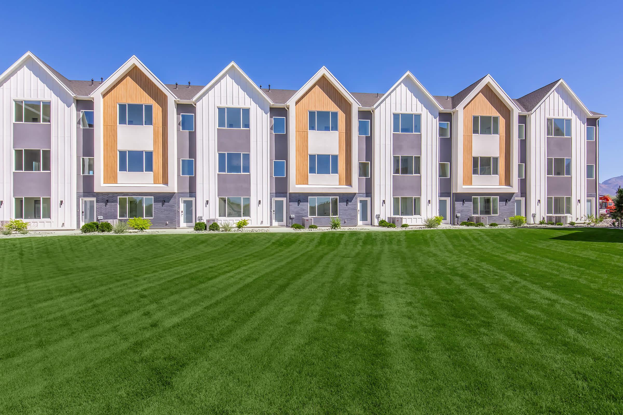 A row of modern townhouses with a mix of white and gray siding, featuring triangular roofs. The buildings have multiple large windows, and the foreground showcases a well-maintained green lawn with neat stripes. Bright blue sky overhead adds to the vibrant atmosphere.
