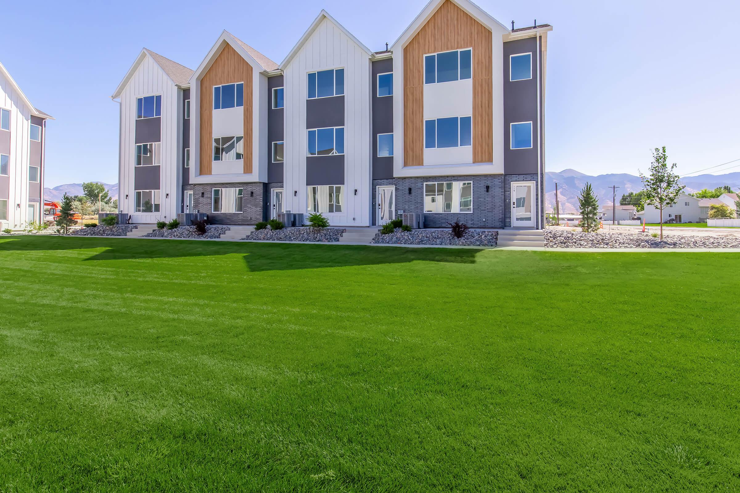 A row of modern townhouses featuring large windows and a mix of gray and wood paneling. The building is set against a bright blue sky and surrounded by a well-manicured green lawn. In the background, mountains can be seen, adding to the scenic view.