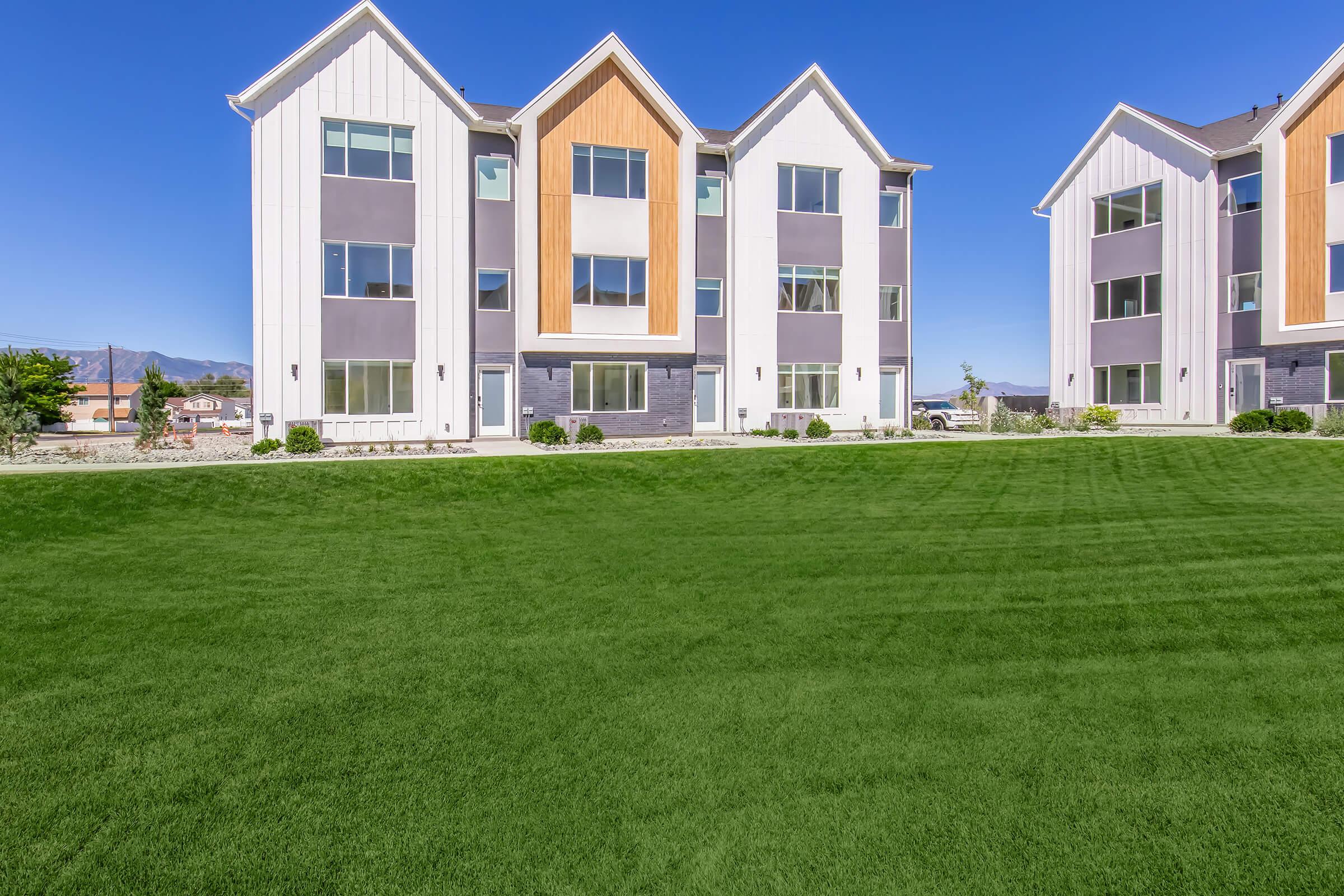 Modern two-story residential buildings with a blend of gray and white siding, featuring large windows. The landscape includes a well-maintained green lawn, with a few shrubs, under a clear blue sky. The buildings have distinct architectural elements, such as angled roofs and wooden accents.