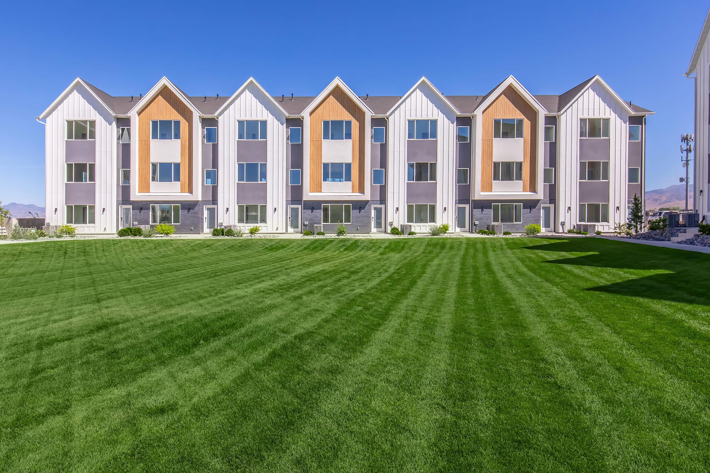 A row of modern townhouses with gabled roofs, featuring a mix of white, gray, and orange facades. The buildings are set against a clear blue sky, with a well-maintained green lawn in the foreground, showcasing neat stripes in the grass.