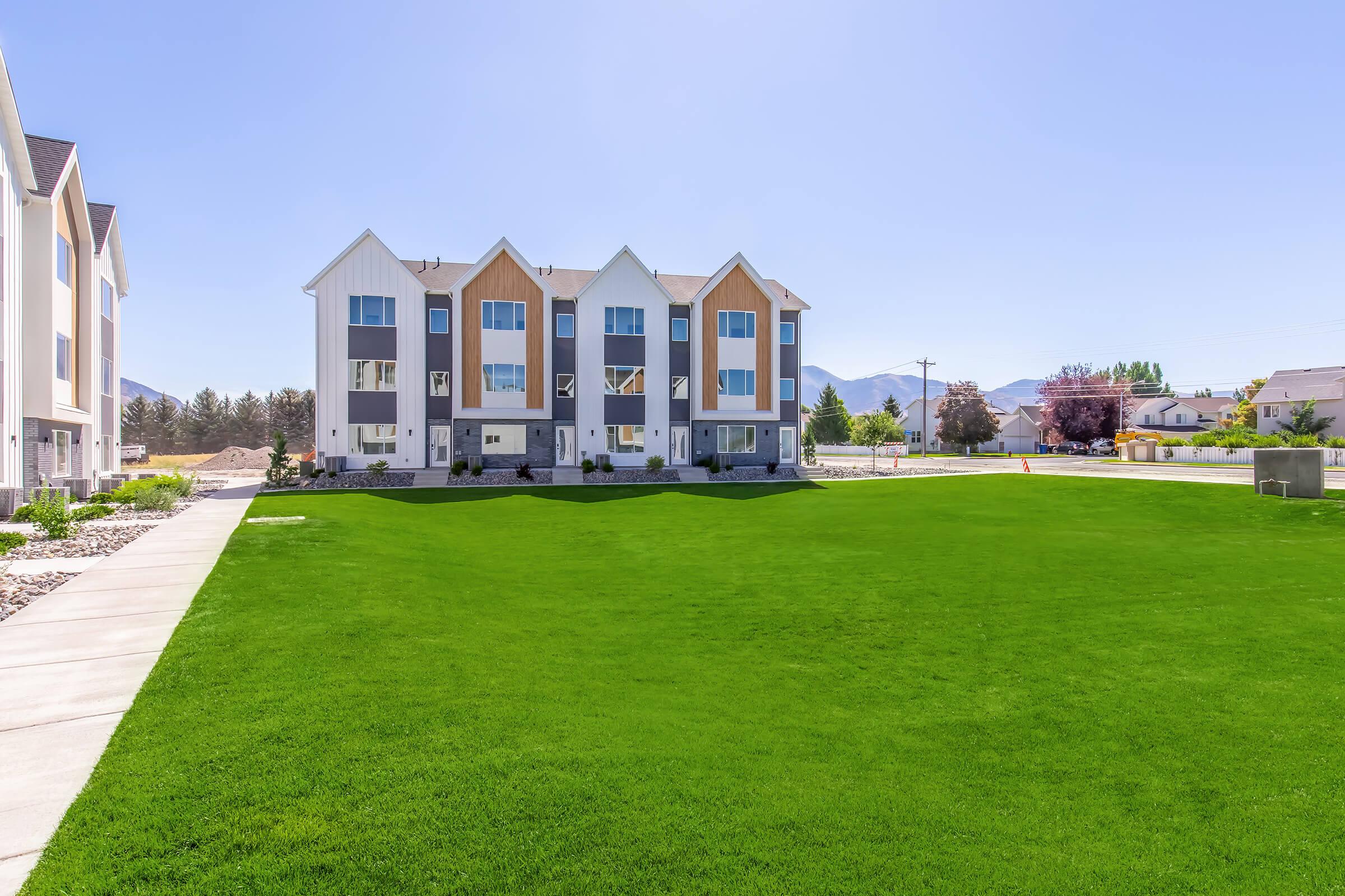 A modern three-story apartment building with a manicured lawn in front. The building features a combination of white and wood paneling, large windows, and a contemporary design. In the background, there are trees and mountains, and the sky is clear. Sidewalks lead to the entrance of the building.