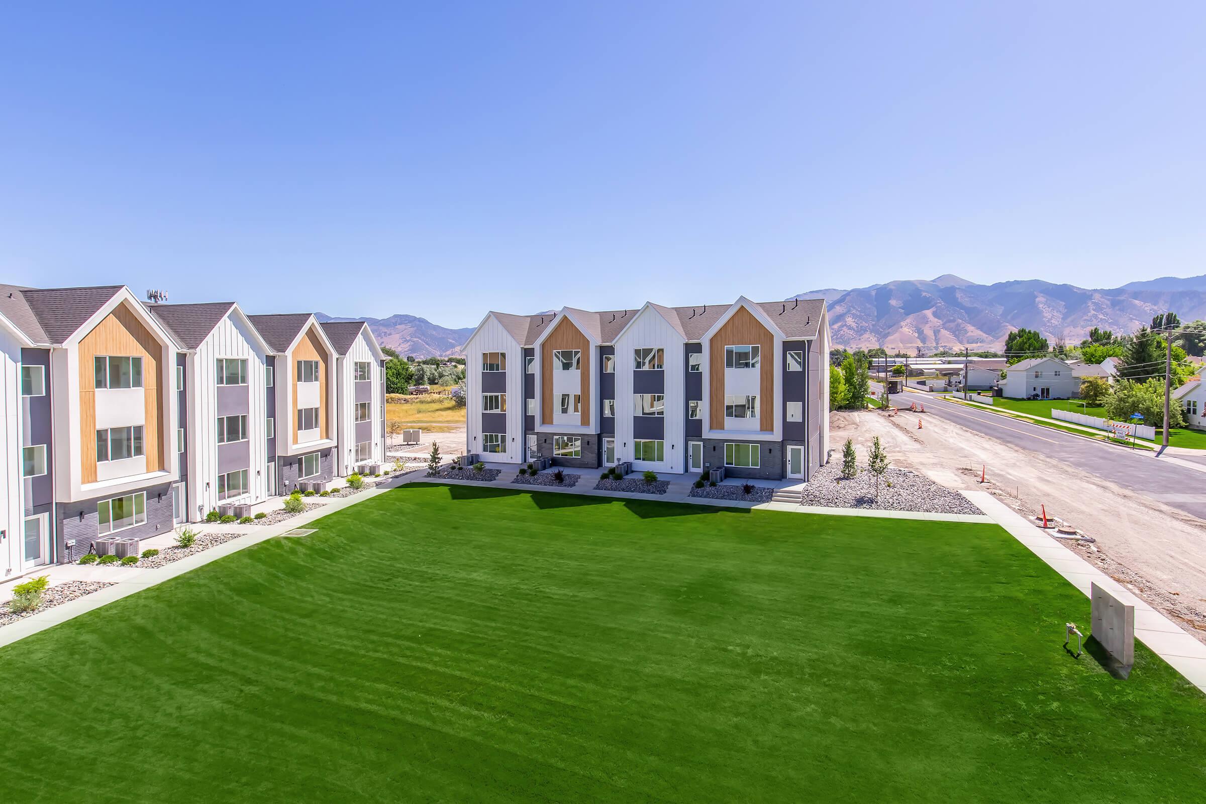 A modern residential building complex with three-story units featuring a variety of colors, set against a backdrop of mountains. The neatly manicured lawn in front highlights a clear blue sky, while a road is visible to the right, indicating a suburban environment.
