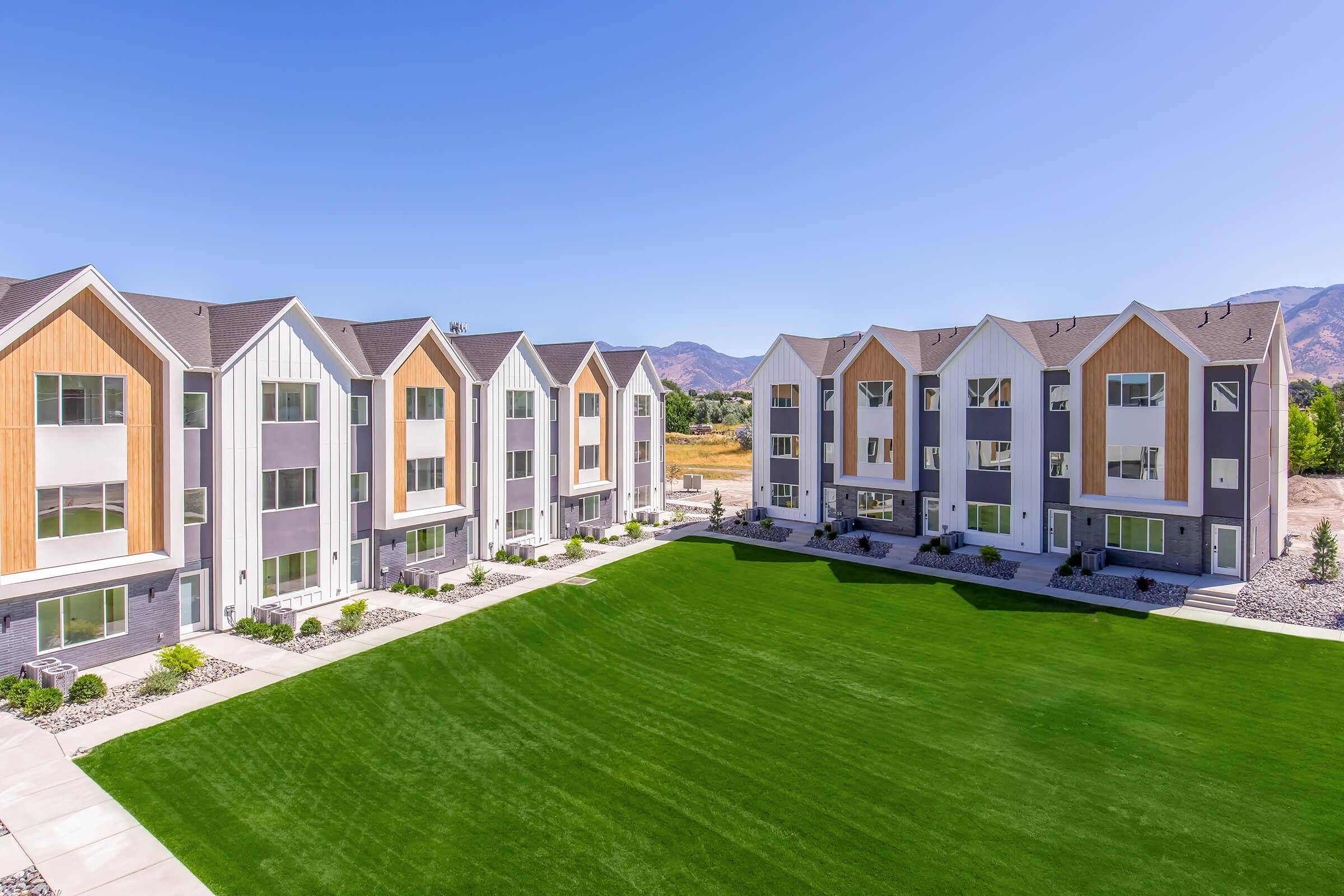 Modern multi-unit residential buildings with a vibrant green lawn in the foreground, set against a clear blue sky and distant mountains. The architecture features a mix of colors and materials, creating an inviting and contemporary living space.