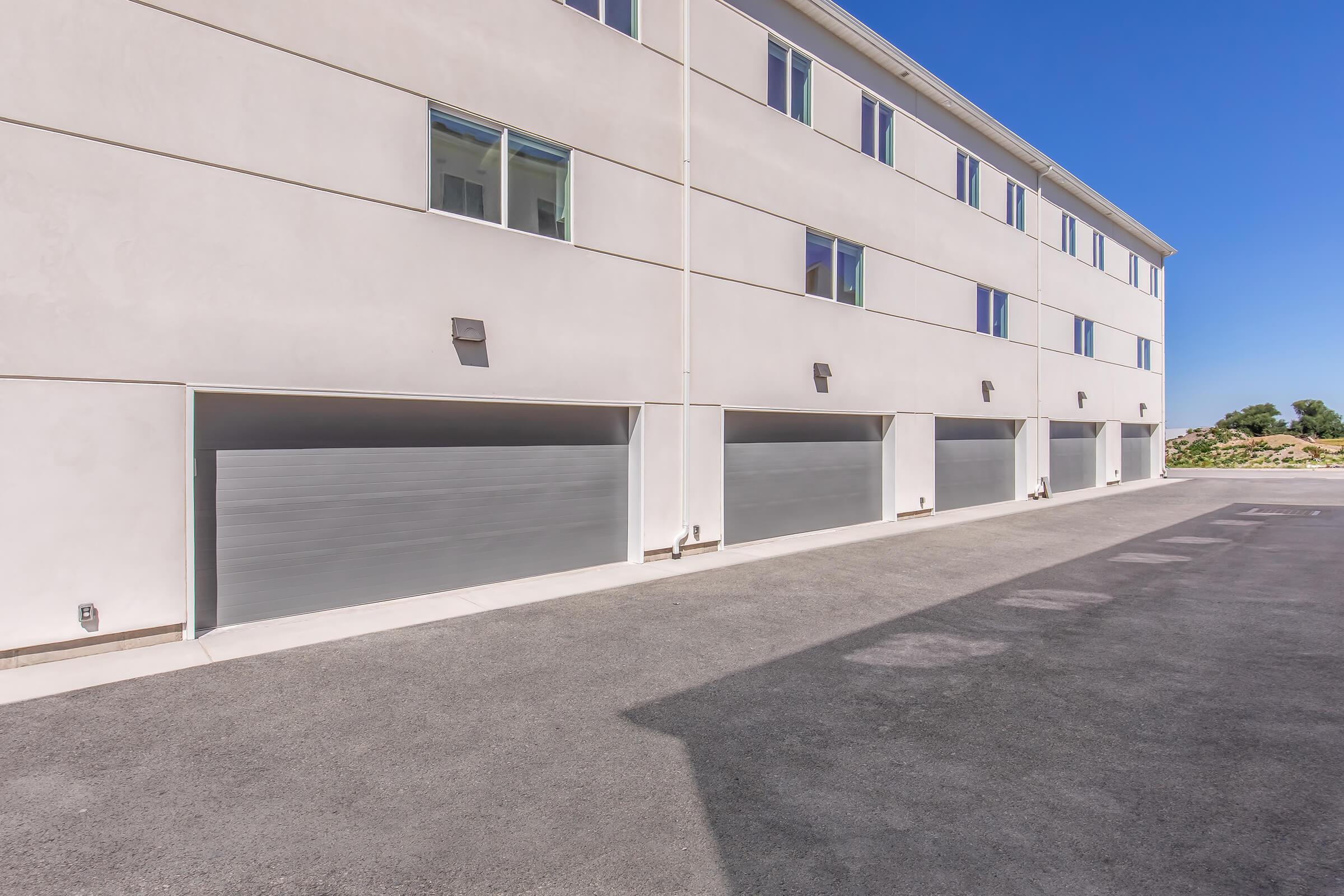A modern building with a smooth, light-colored exterior featuring multiple garages. The garages have sleek, metallic doors and are set against a clear blue sky. A paved area is visible in front of the garages.