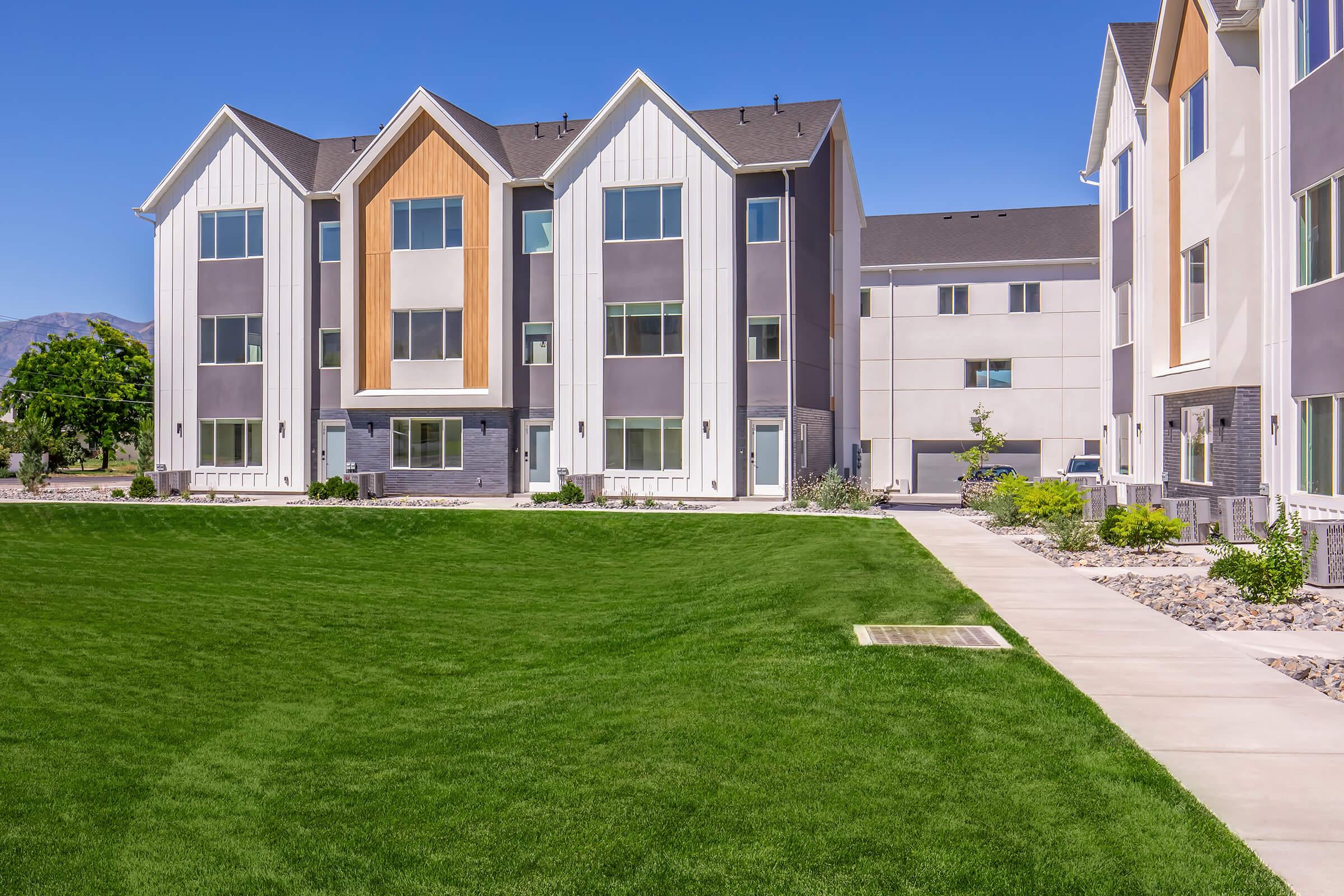Modern apartment building featuring a combination of grey, white, and wood paneling. The landscape includes well-maintained green lawns and a paved walkway. Bright blue skies are visible in the background, enhancing the fresh and inviting atmosphere of the property.