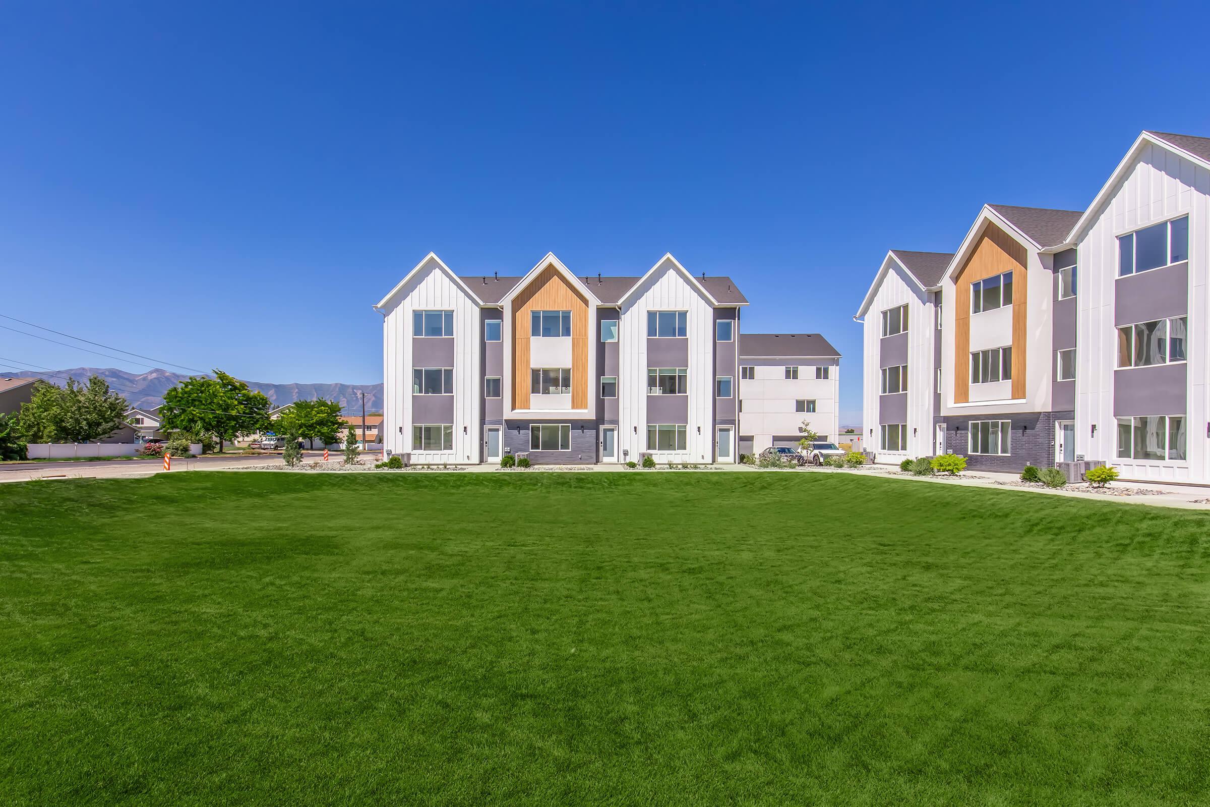 A modern residential complex featuring multiple three-story buildings with a mix of colors, surrounded by a vibrant green lawn. Clear blue sky and distant mountains are visible in the background, creating a serene atmosphere. Ideal for outdoor activities and community living.