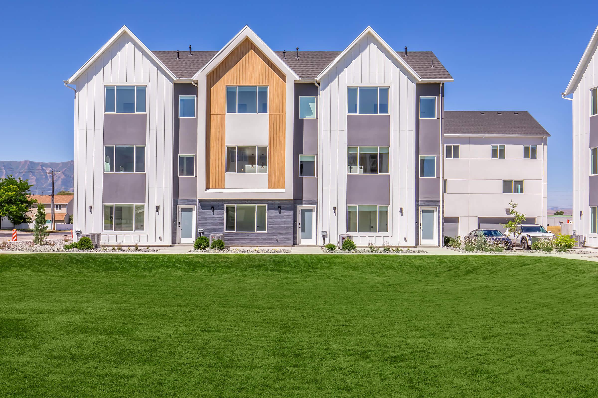 A modern multi-unit residential building with a combination of white, gray, and wood paneling. The three-story structure features multiple large windows, a green lawn in front, and surrounding landscaping. Clear blue sky in the background.