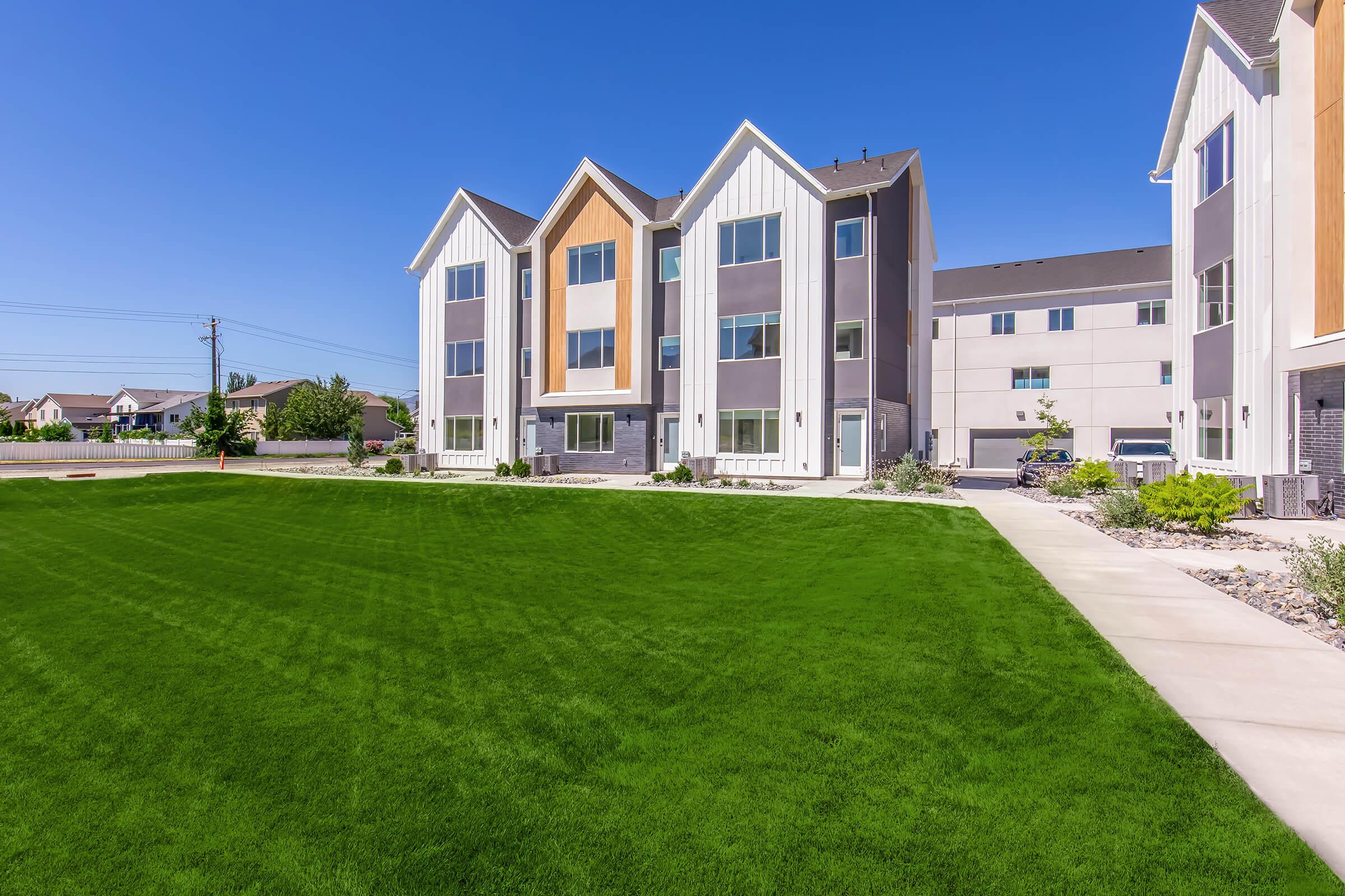 Modern multi-family residential building with a combination of gray, white, and wooden facades. The structure features three prominent gable roofs and large windows. A well-maintained green lawn surrounds the building, and a paved pathway leads to the entrance under a clear blue sky.