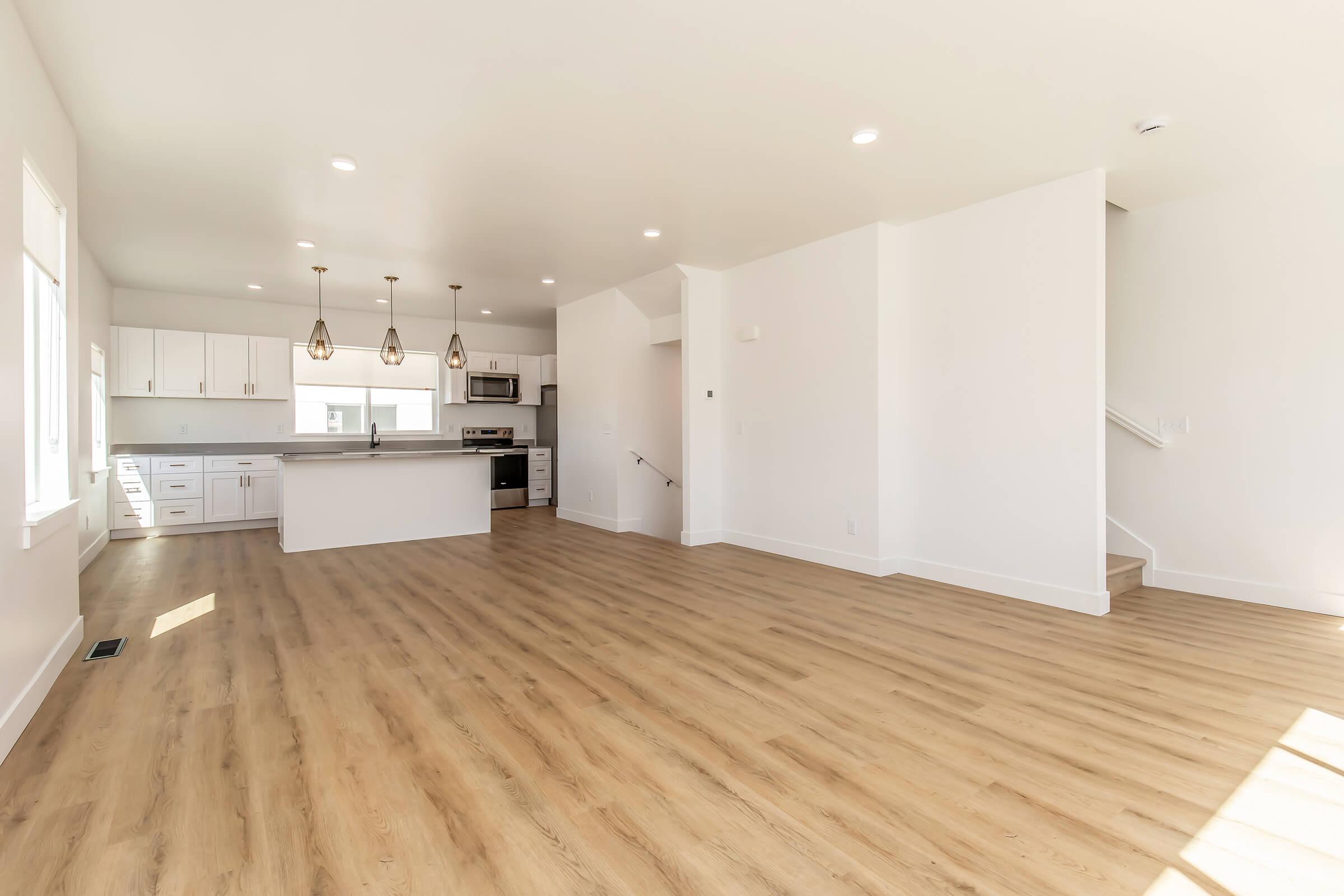 Spacious, empty interior of a modern living area featuring light hardwood floors, white walls, and large windows. A kitchen area is visible with pendant lighting, and there is an open layout leading to a staircase in the background. Natural light illuminates the space, emphasizing its airy feel.