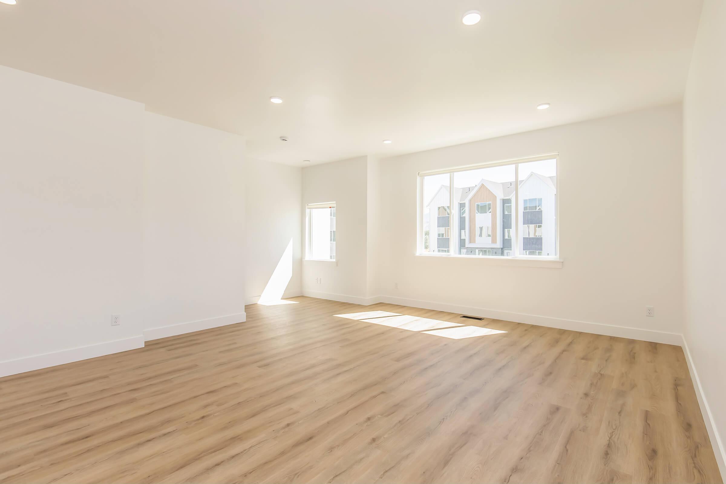 A spacious, empty room with white walls and light-colored wooden flooring. Large windows allow natural light to fill the space, casting shadows on the floor. The room is bare, suggesting it may be ready for furniture or decoration, with clean lines and a modern design.