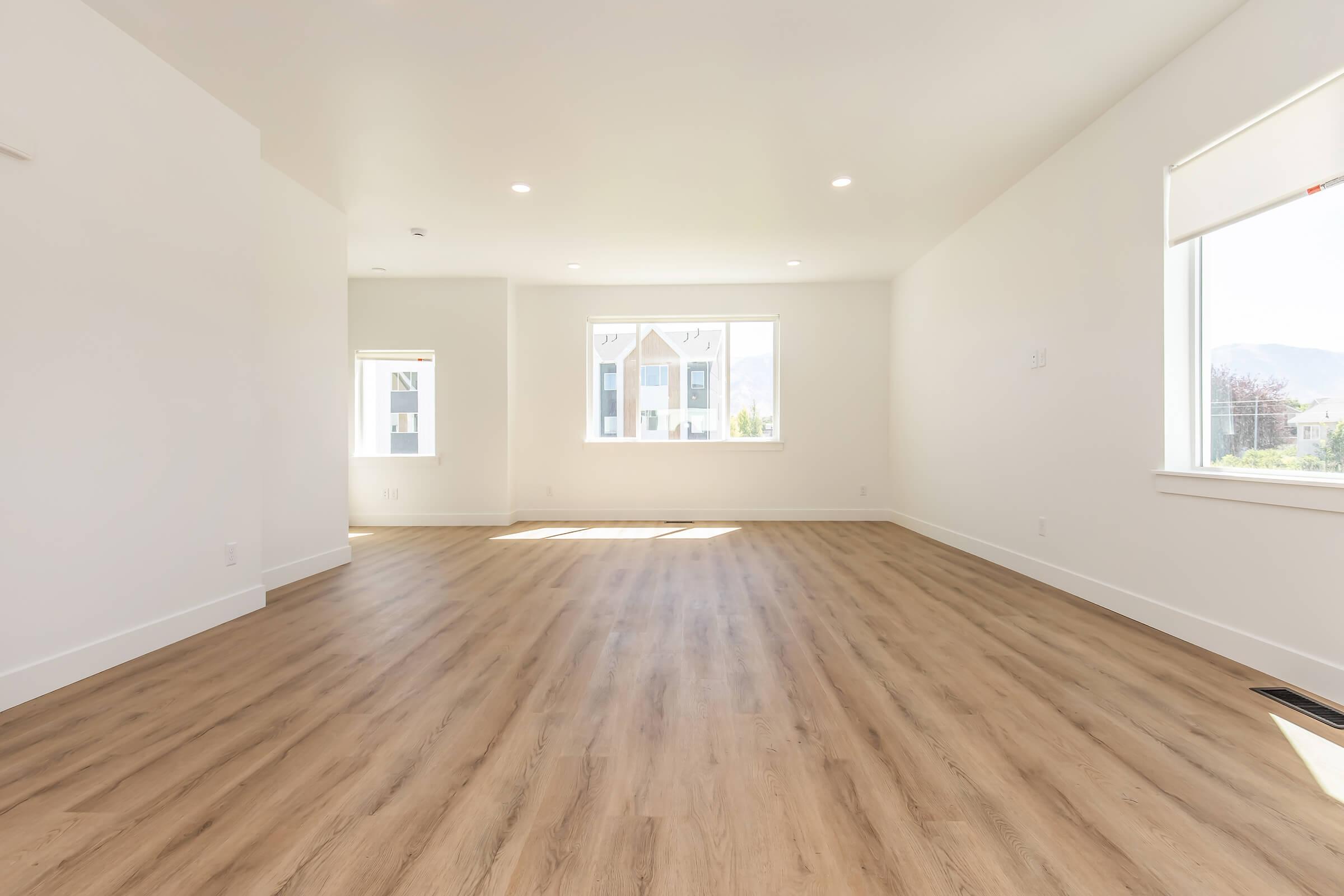A spacious, brightly lit room with white walls and large windows allowing natural light. The floor is covered with light-colored wood laminate, creating a warm atmosphere. The room is empty, showcasing its open layout, with another window visible in the background.
