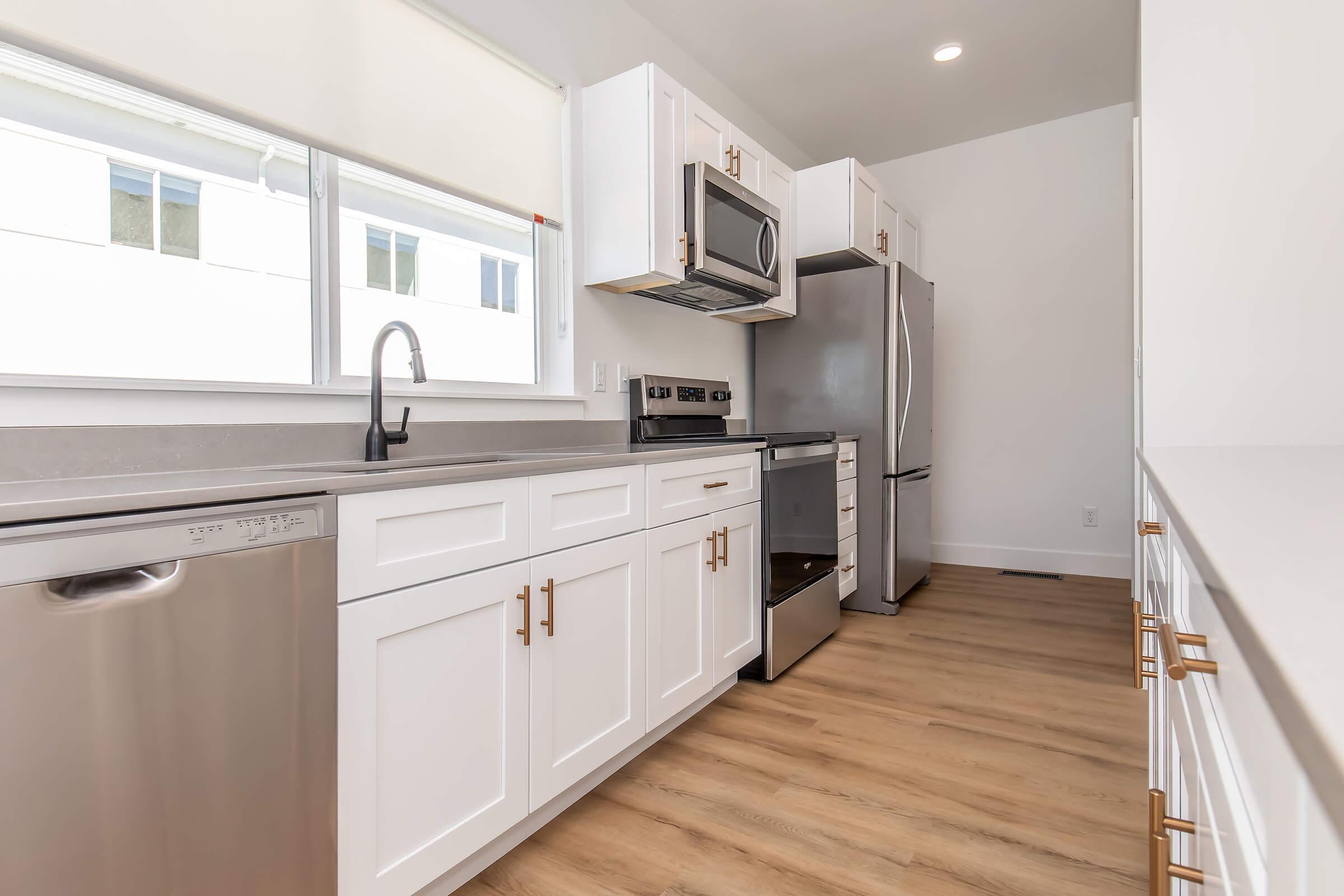 Modern kitchen featuring white cabinets with gold hardware, stainless steel appliances including a refrigerator and dishwasher, and a large window allowing natural light. The flooring is light wood, creating a warm and inviting atmosphere.