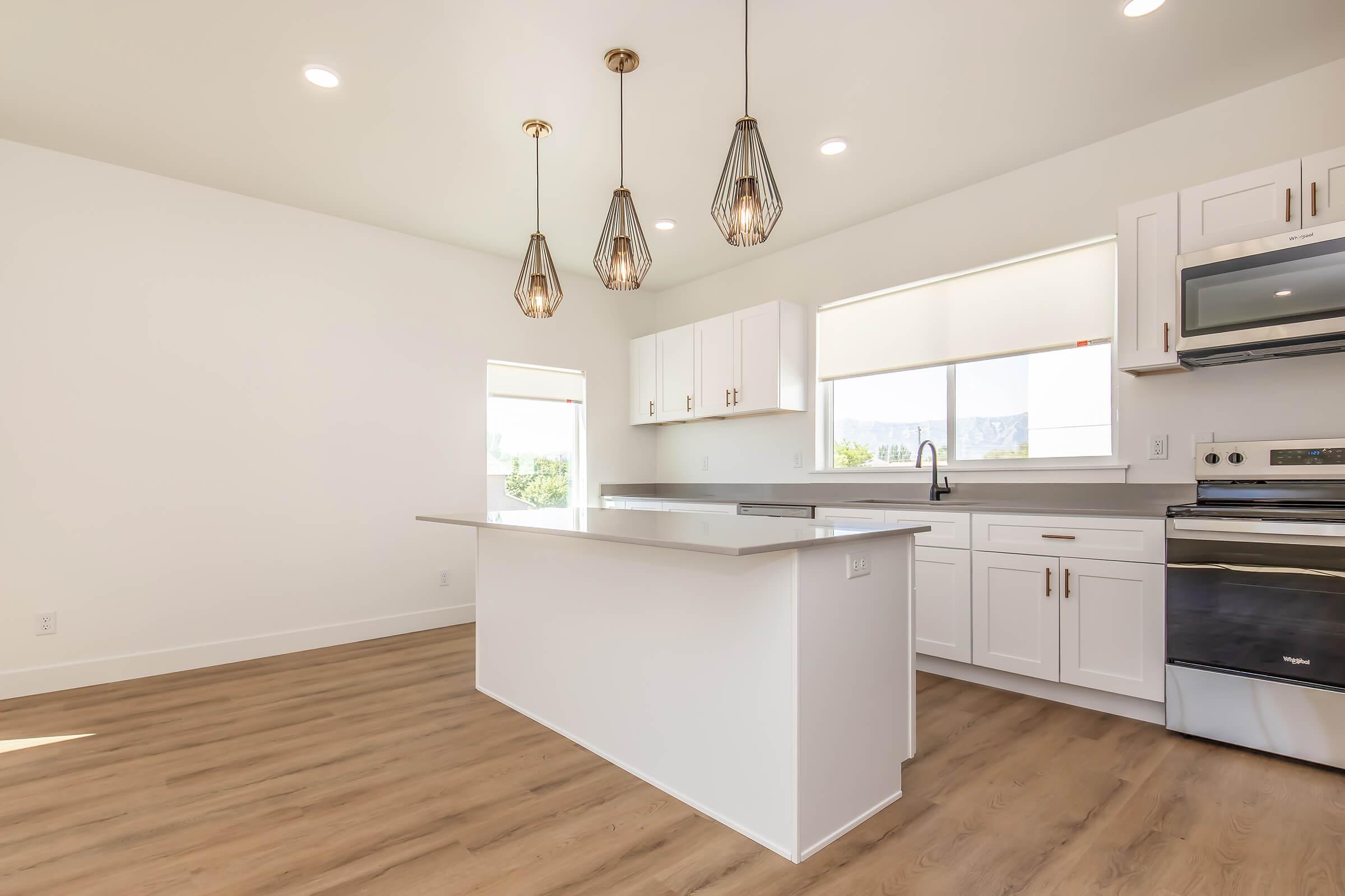Modern kitchen with white cabinets, stainless steel appliances, and a large island. Three pendant lights hang over the island, and there is a window providing natural light. The flooring is light wood, and the walls are painted white, creating a clean and bright ambiance.