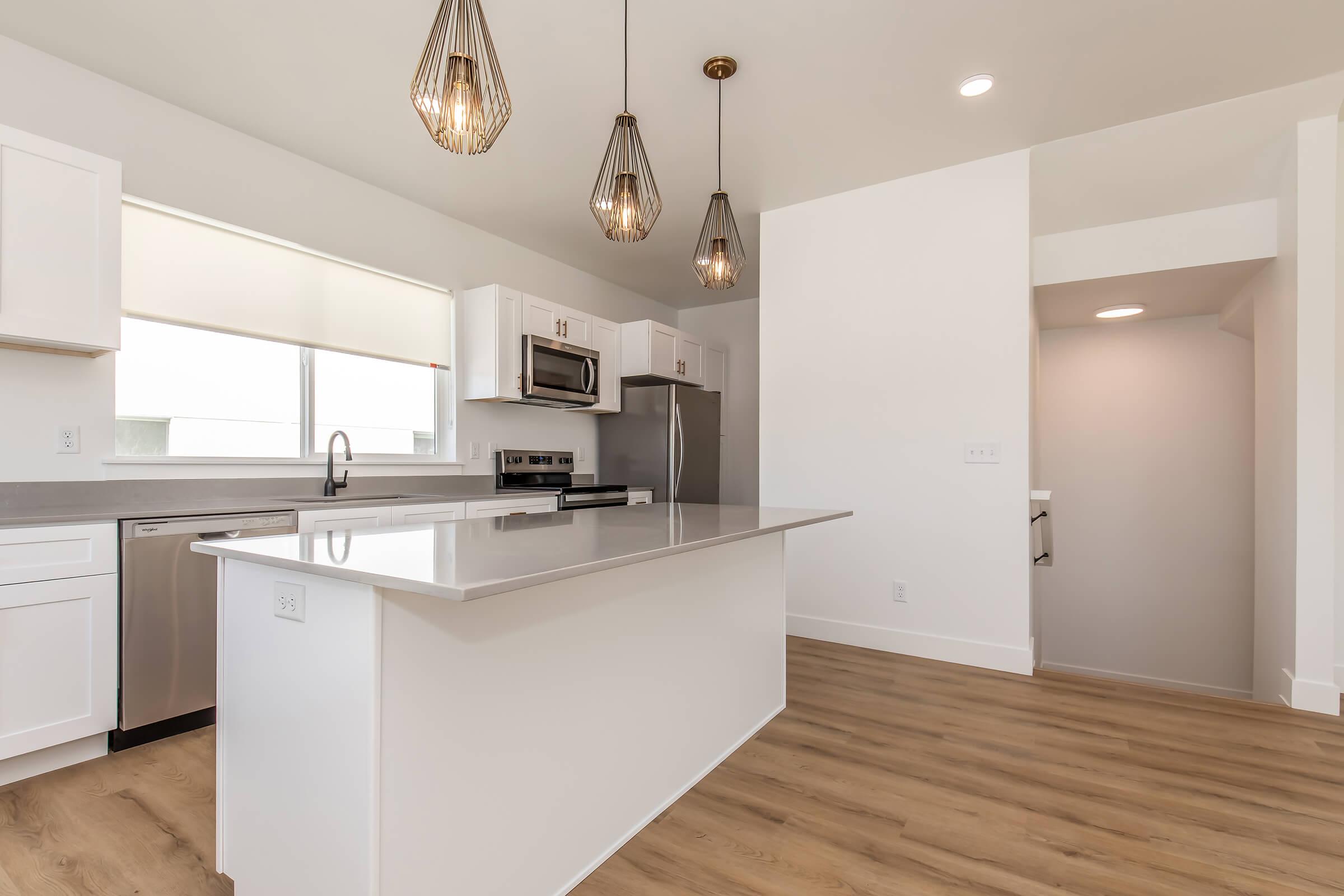 Modern kitchen featuring a central island with a white countertop, stainless steel appliances, and stylish pendant lighting. The space has light wood flooring, white cabinetry, and a large window that provides natural light. An open layout connects to an adjacent room.