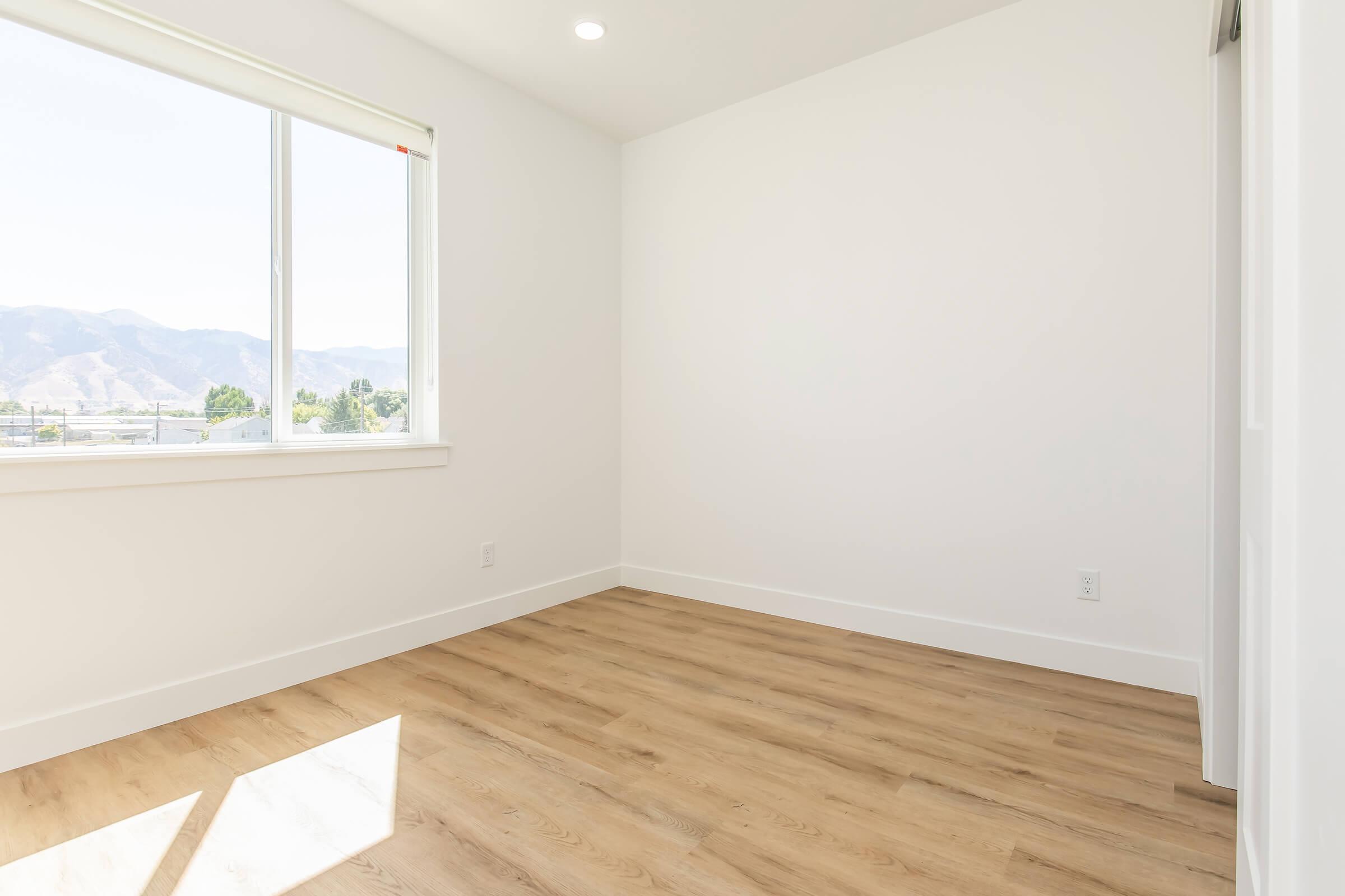 A bright, empty room with white walls and a large window letting in natural light. The floor is made of light-colored wood, and in the background, distant mountains are visible through the window. The space appears modern and minimalistic, perfect for personal decoration.