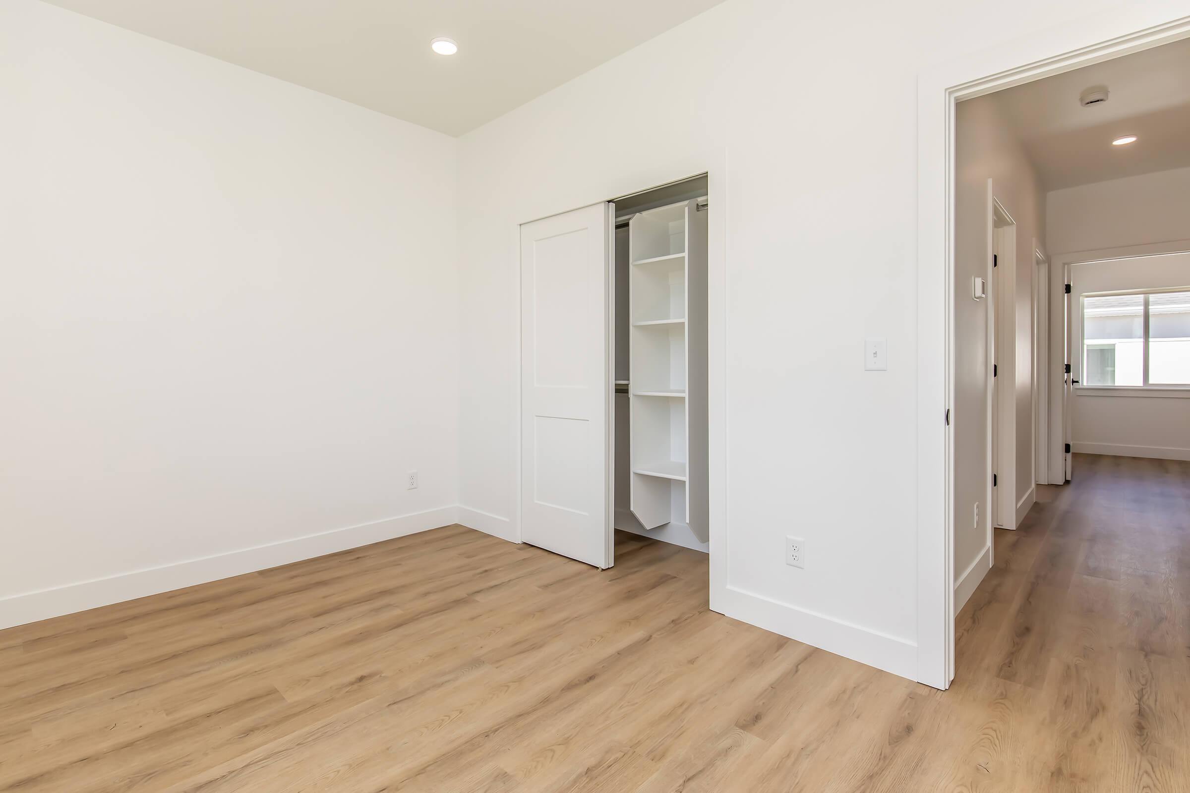 A modern, empty room with light-colored walls and hardwood flooring. There is a closet with sliding doors on the left, and a doorway leading to another room in the background. Natural light enters from a window out of view.