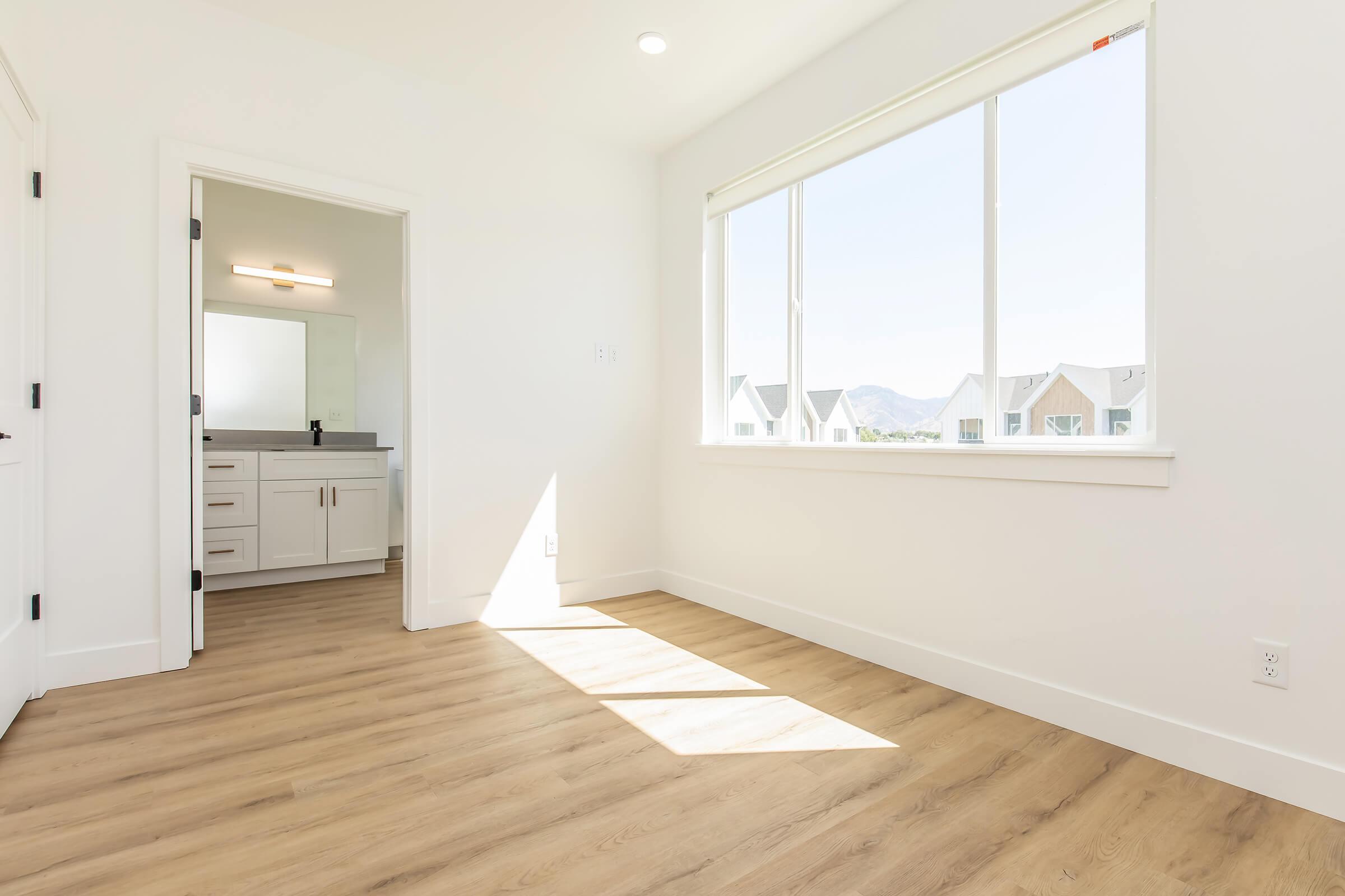 Bright and airy interior space featuring a large window that lets in natural light, with light-colored walls and modern wooden flooring. On the left, a doorway leads to a stylish bathroom with a mirror and sink. In the background, distant hills are visible through the window.