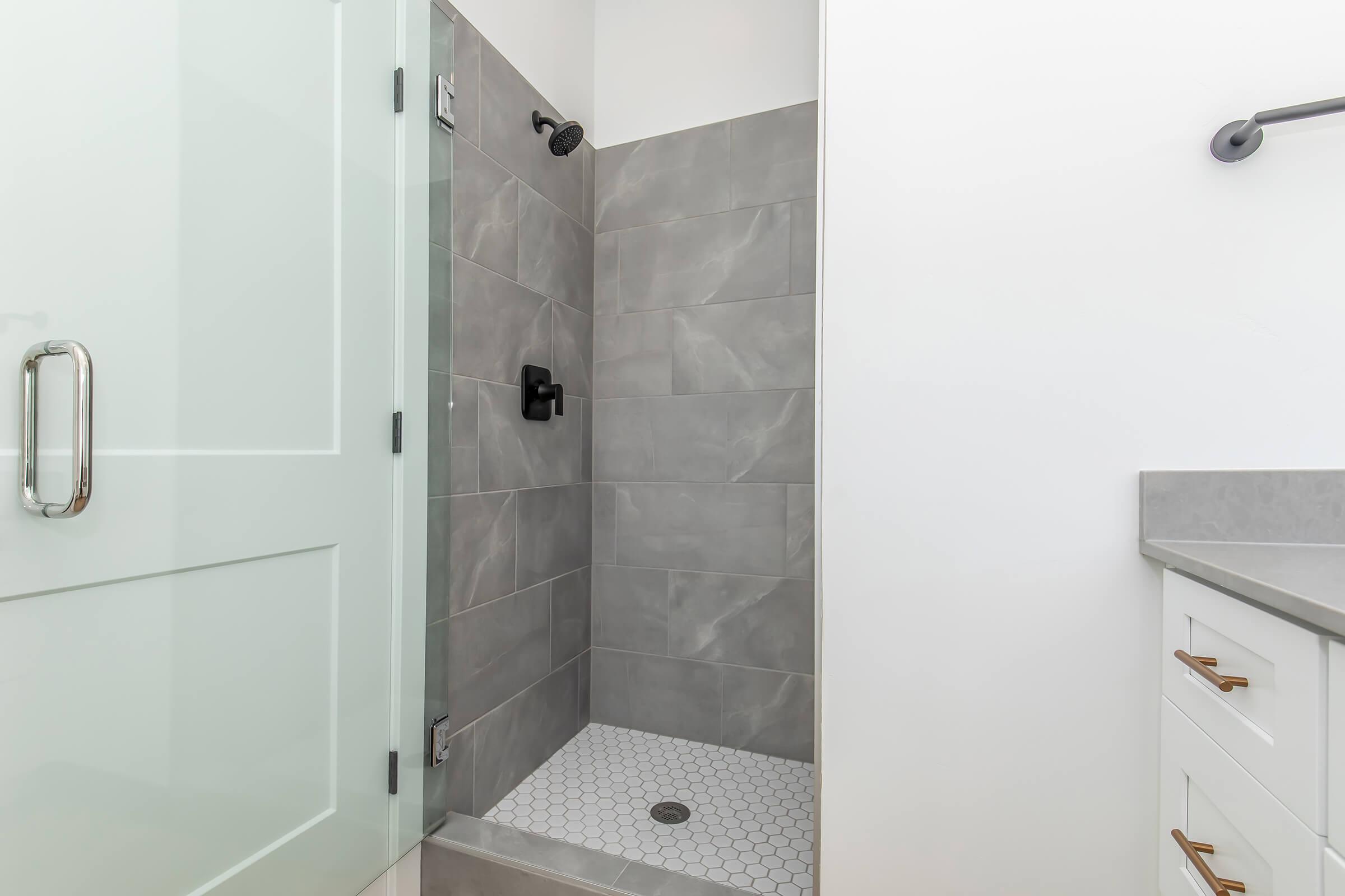 Modern shower area featuring a glass door, gray tiled walls, and a hexagonal tiled floor. A sleek black showerhead and fixtures are visible. The bathroom also includes a countertop with a sink and cabinet, enhancing a contemporary aesthetic. Overall, the space is bright and clean.