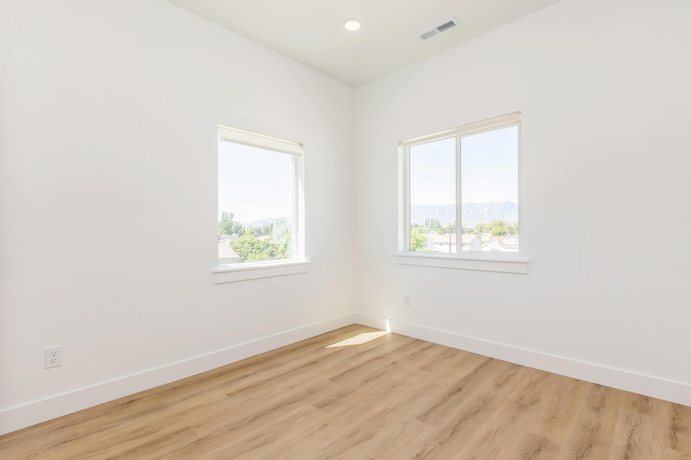 A bright, empty room with two windows offering a view of mountains. The walls are white, and the flooring is light-colored wood. Natural light floods the space, highlighting the minimalist design and spacious feel.
