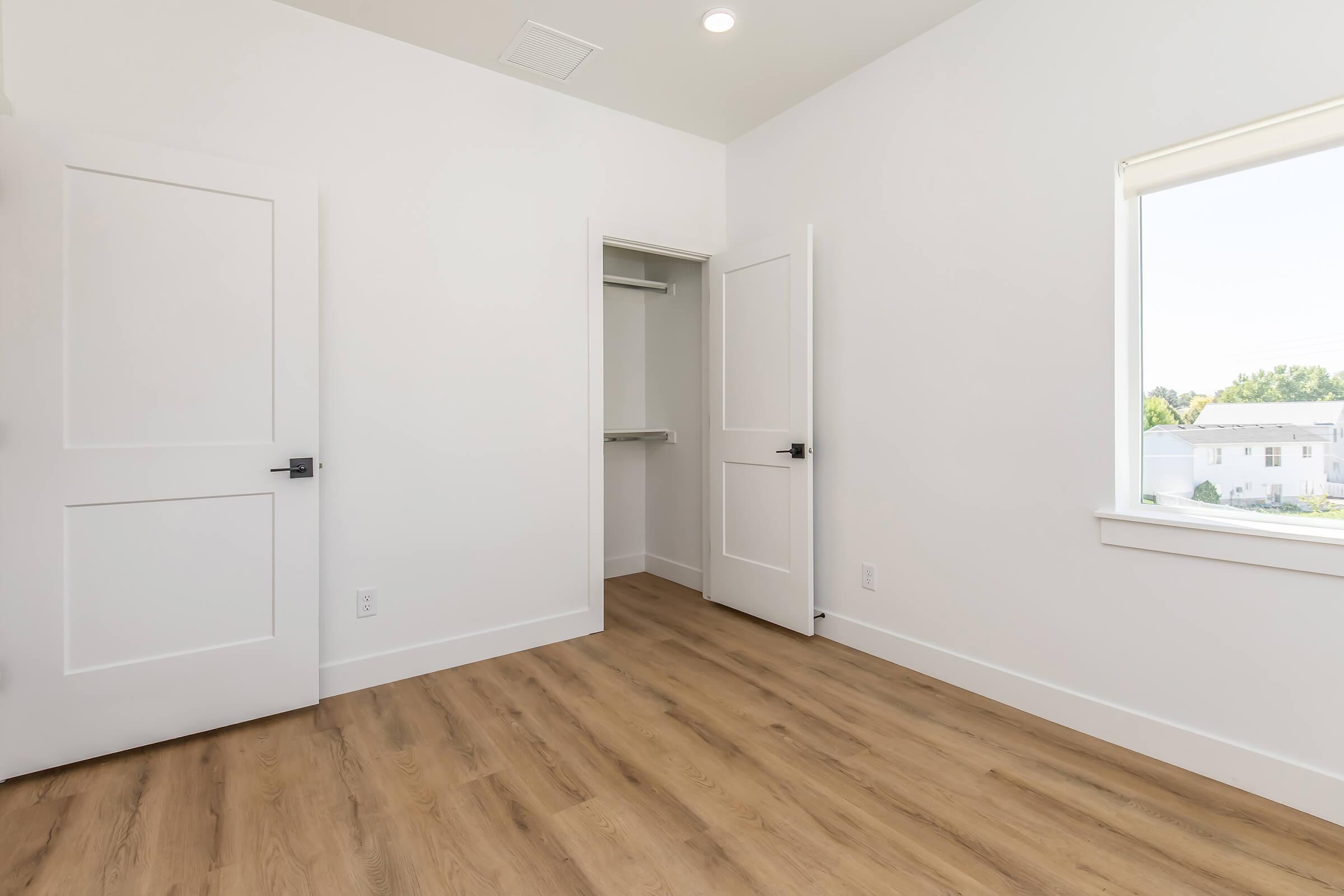 A bright, empty room featuring light-colored walls, wood-style flooring, and two closed white doors. One door leads to a closet with a small opening, and a window on the far wall allows natural light to enter, creating a spacious and minimalist atmosphere.