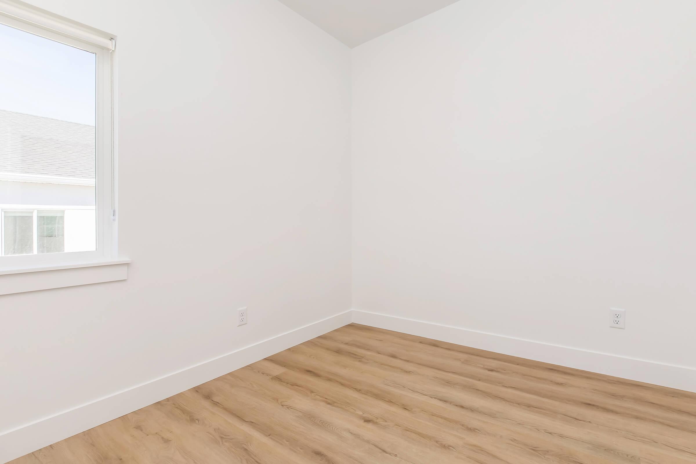 A minimalistic room with white walls and light wooden flooring. A window lets in natural light, illuminating the empty space, which has no furniture or decorations, creating a clean and airy atmosphere.