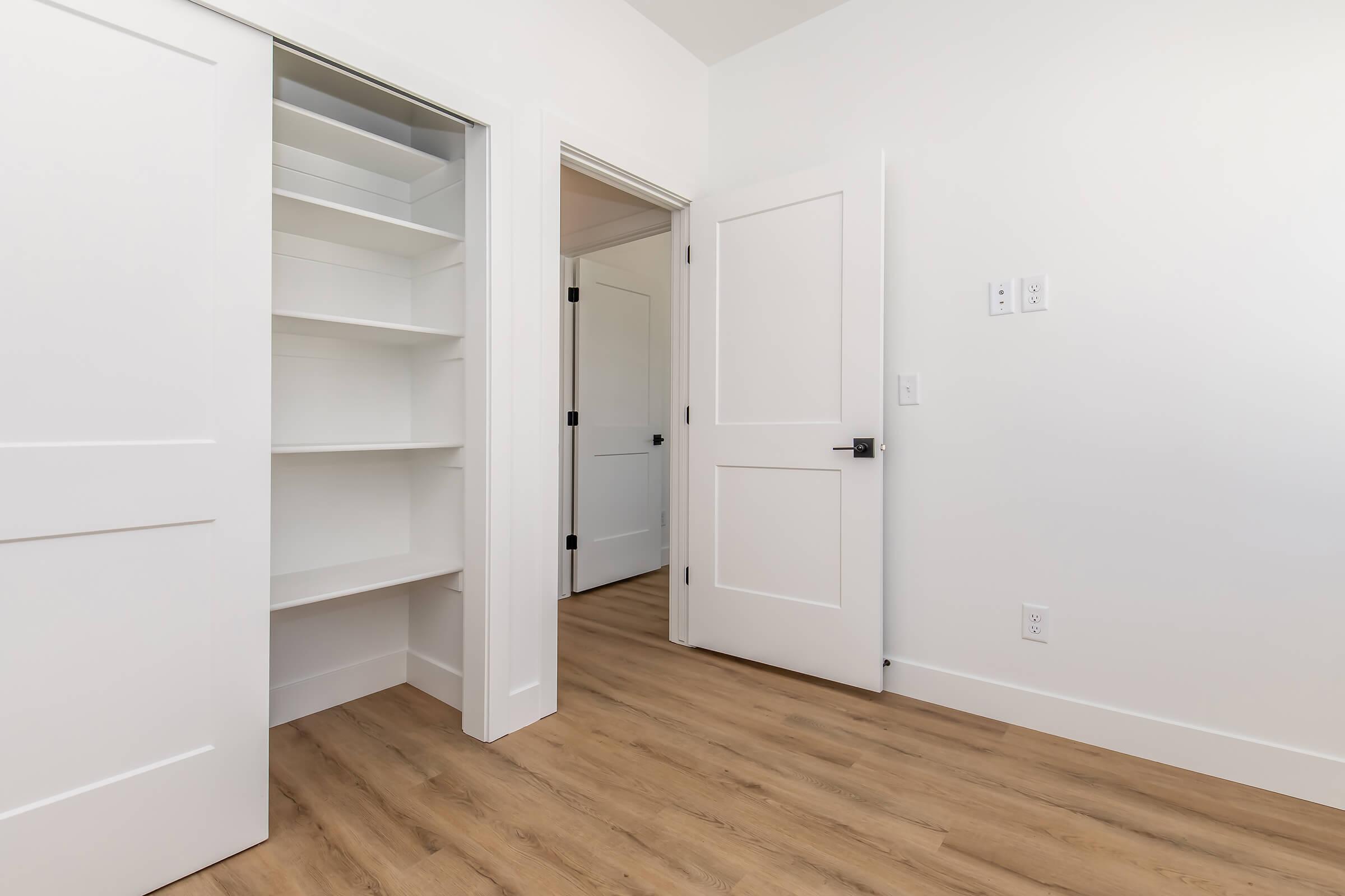 Interior view of a room featuring a doorway leading to a closet with shelves, a second door to another area, and light wood flooring. The walls are painted white, creating a bright and clean aesthetic.