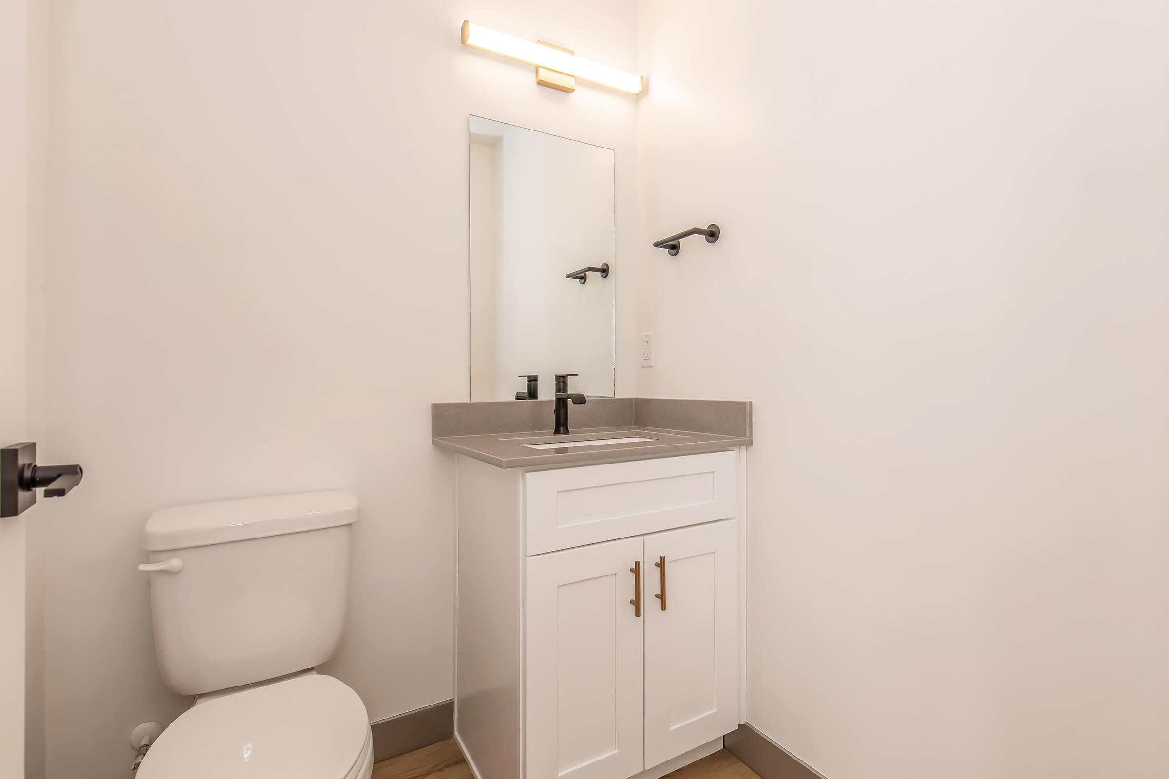 Small modern bathroom featuring a white toilet, a sleek vanity with a gray countertop, and a mirror illuminated by a wall-mounted light fixture. The walls are painted white, and the flooring is light wood.