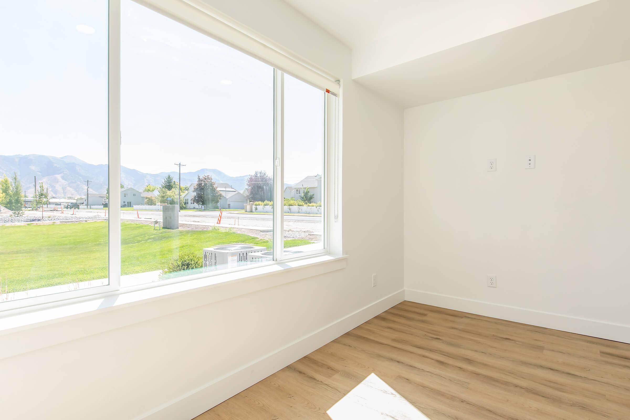 Bright and airy room with large windows overlooking a green landscape and distant mountains. The walls are white, and the floor features light wood accents. Natural light streams in, creating a welcoming atmosphere. The scene suggests a tranquil and modern living space.