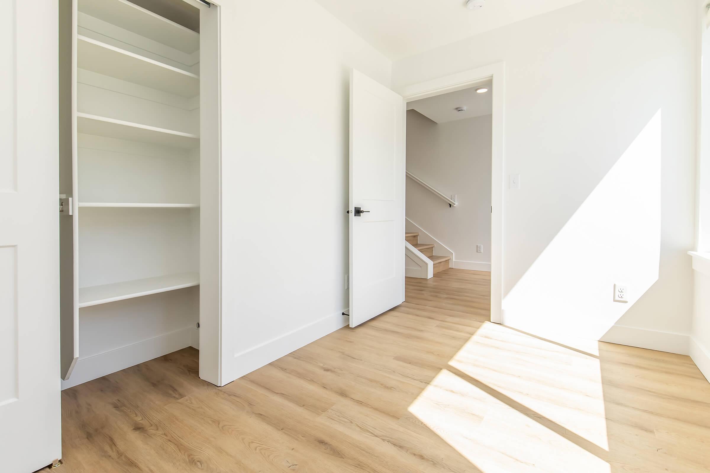 A bright, empty room with light-colored walls and wooden flooring. There is an open closet with shelves on the left and an open door leading to stairs on the right. Natural light streams in through a window, casting shadows on the floor.