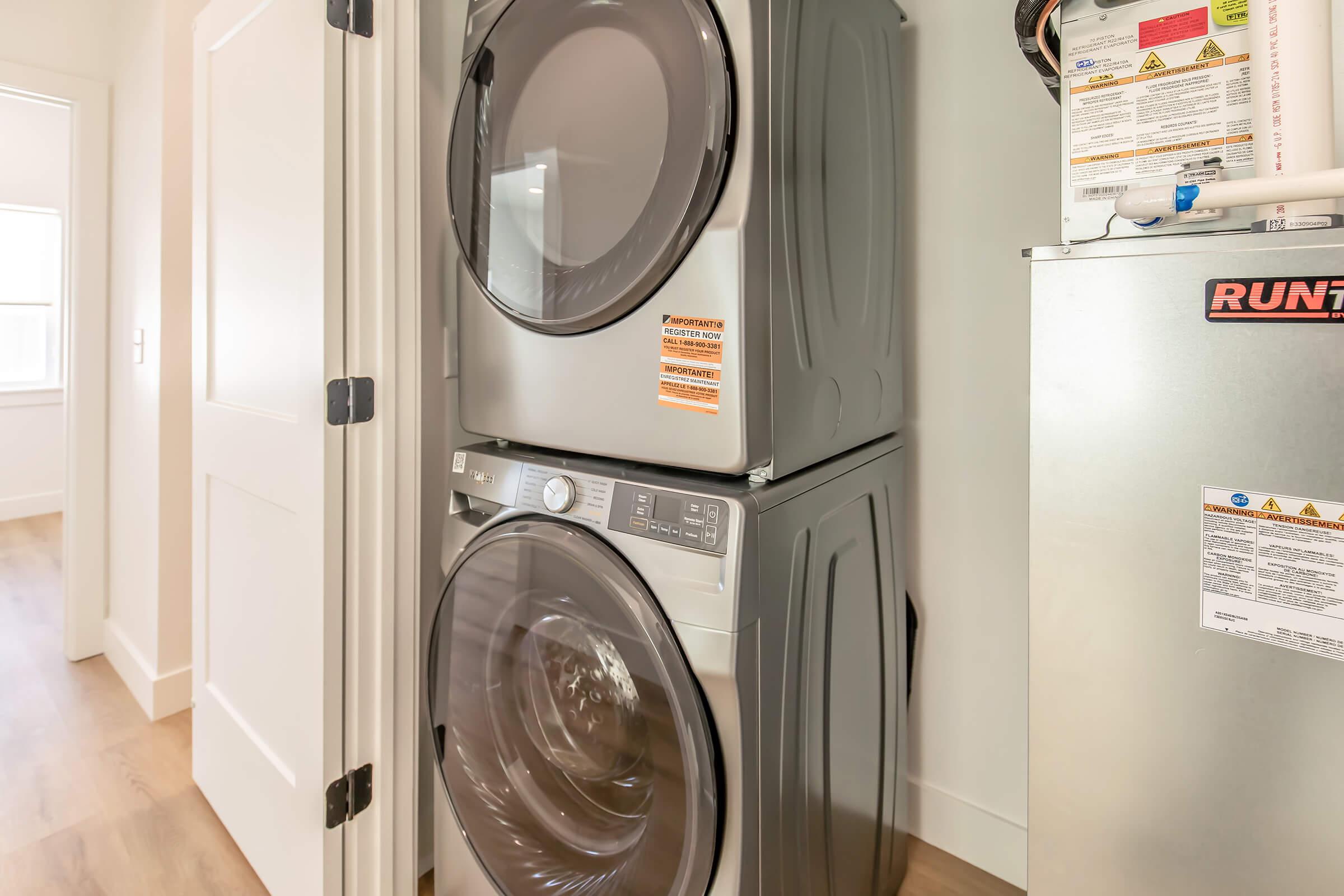 A stackable washer and dryer in a laundry room. The appliances are silver with front-loading doors. There's a door leading to another room in the background, and a metal appliance, likely a dryer or utility unit, is visible beside them. The floor is light-colored wood. The walls are painted white.
