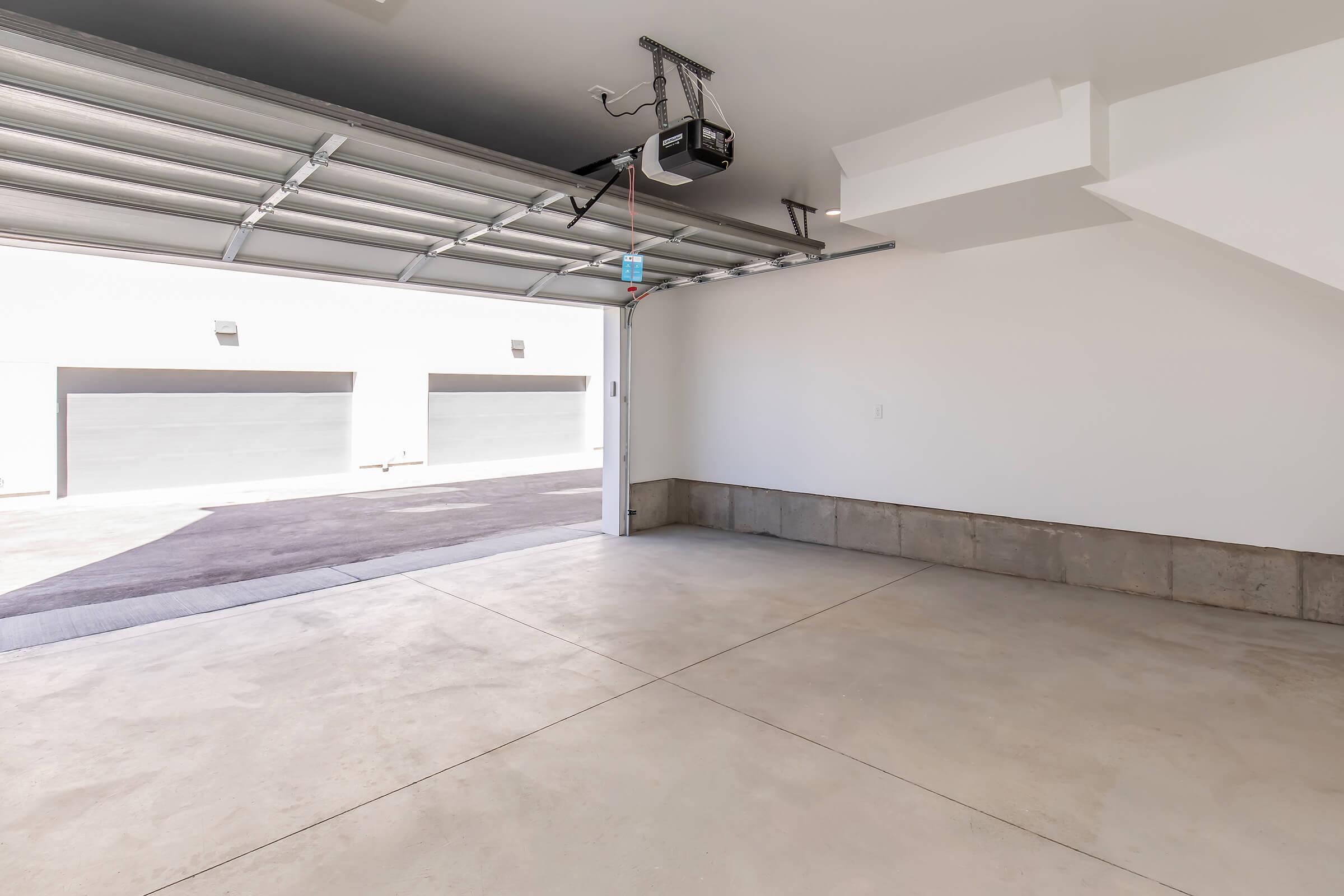 Interior of a spacious, empty garage with a concrete floor. The garage door is open, revealing a view of multiple closed garage doors outside. The walls are plain and painted white, with a ceiling-mounted garage door opener visible. The overall atmosphere is clean and bright.