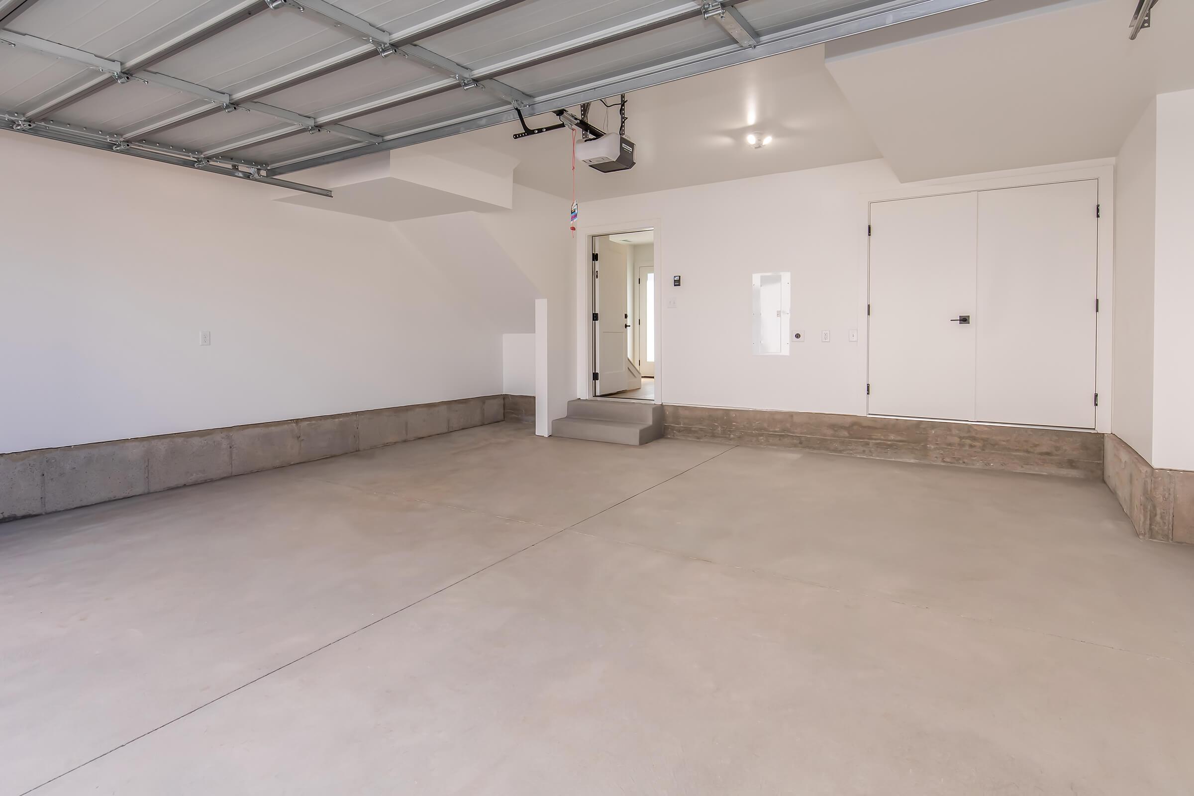 A clean, empty garage with concrete flooring, white walls, and a high ceiling. There is a closed door on the right leading to another room, and stairs on the left leading to an upper level. The garage is well-lit with overhead lighting and a pull-down garage door.