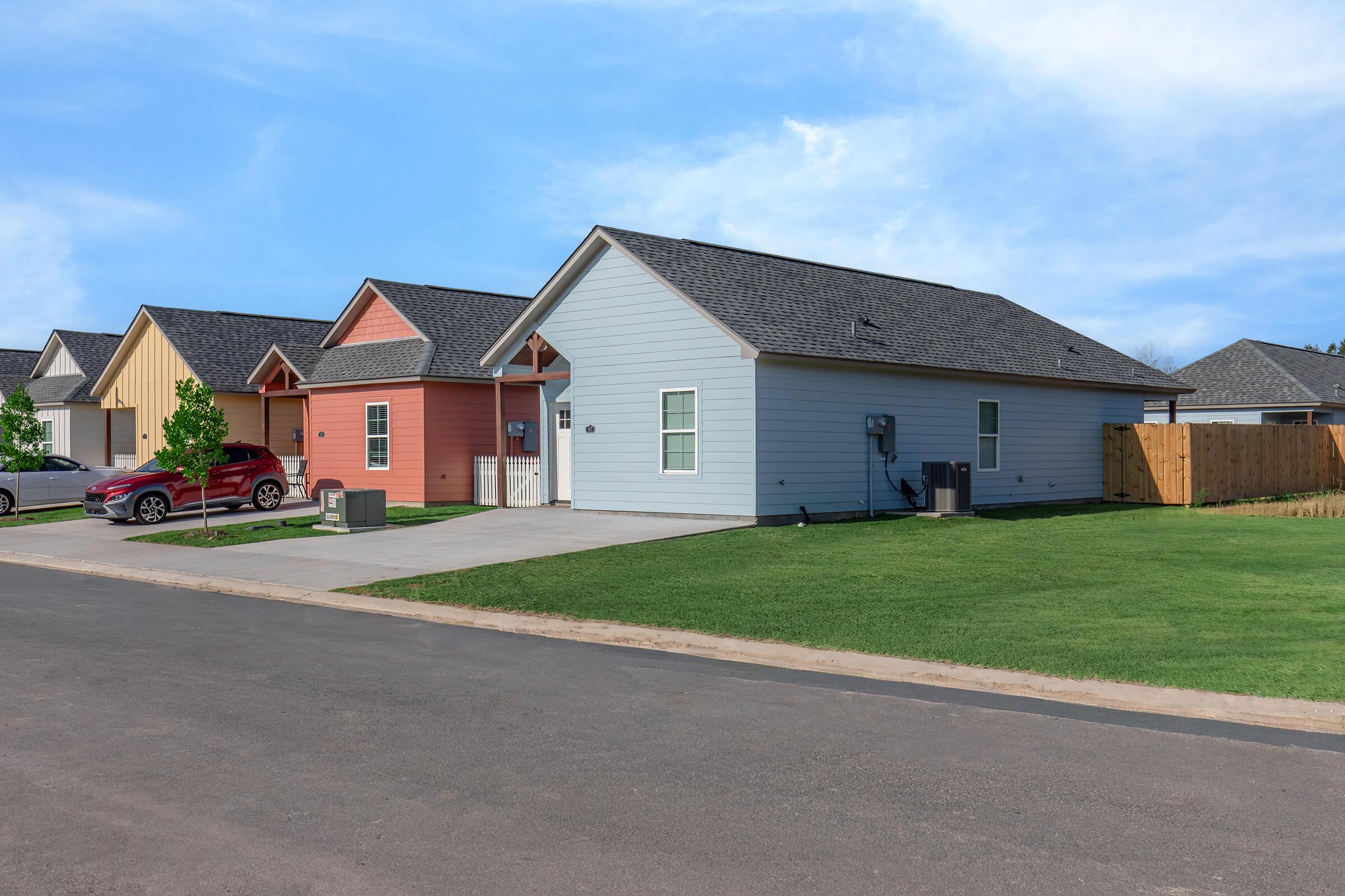 A residential street featuring two houses, one in light blue and the other in peach, both with gable roofs. The houses are surrounded by green lawns, with a paved driveway in front of them. A red car is parked in front of the light blue house, and a wooden fence is visible in the background.