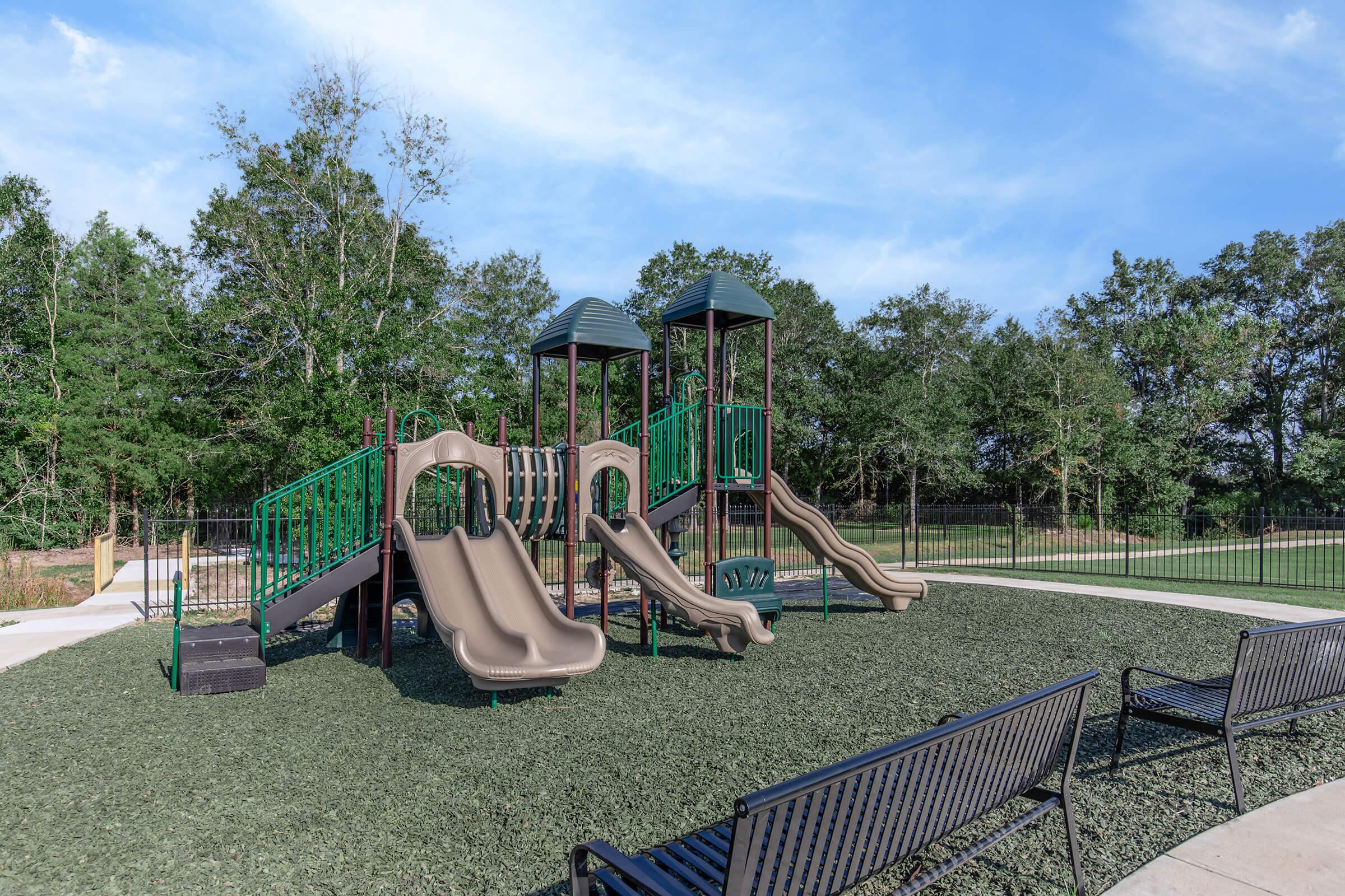 A playground featuring two beige slides and green climbing structures surrounded by artificial grass. In the foreground, there are black benches along a pathway, with trees and a blue sky in the background. The area is enclosed by a black fence.