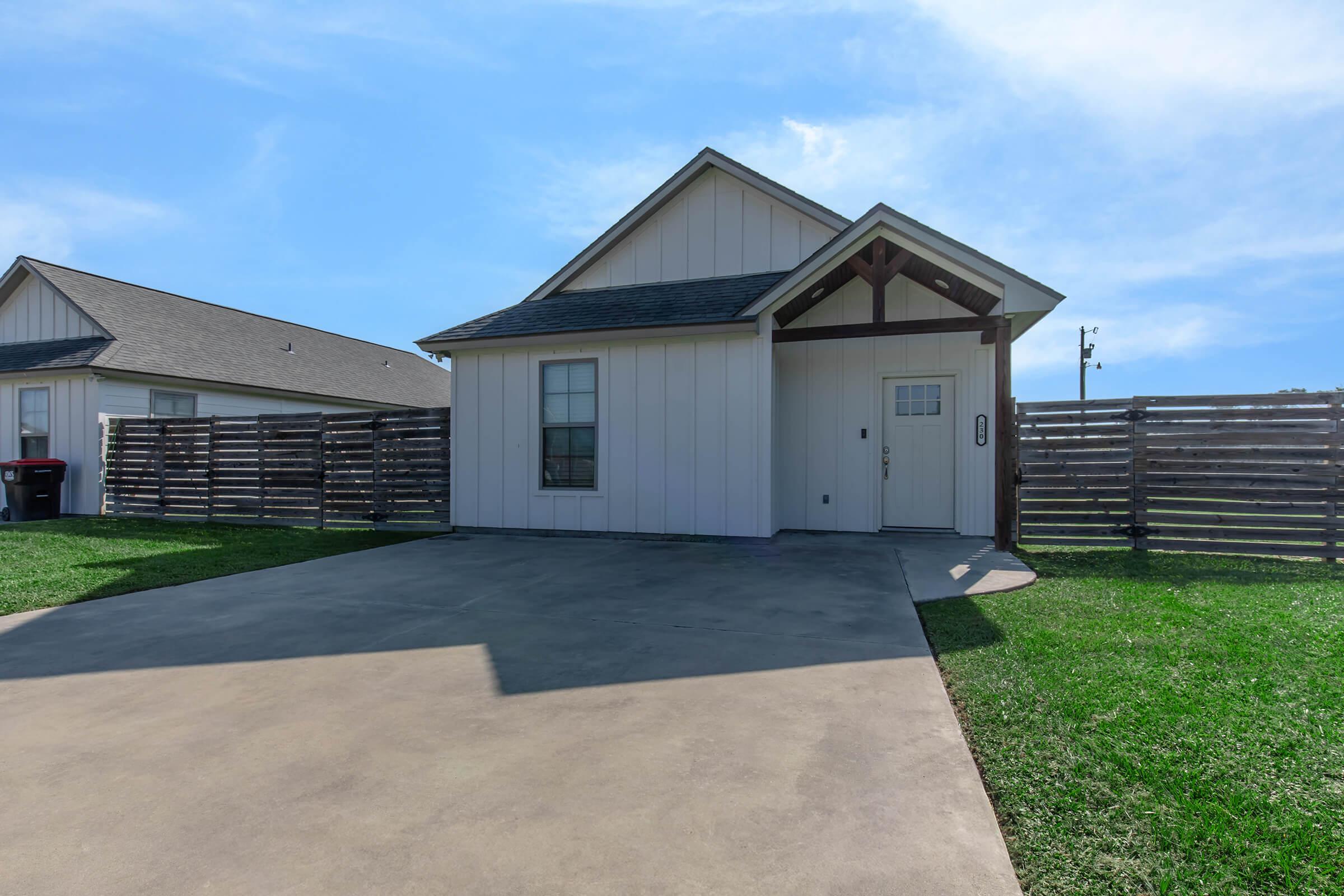 A small, modern house with a peaked roof and a light-colored exterior. The front features a single door and a window. A concrete driveway leads up to the house, which is surrounded by a wooden fence. Lush green grass is visible in the front yard, and a nearby house can be seen on the left side.
