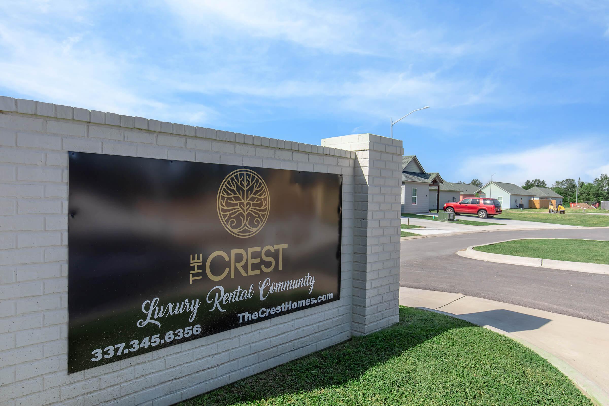 A promotional sign for "The Crest Luxury Rental Community" featuring a phone number and website. The sign is mounted on a brick wall, with a clear view of a residential area in the background, including newly constructed homes and a red vehicle on the street. The sky is blue with some clouds.