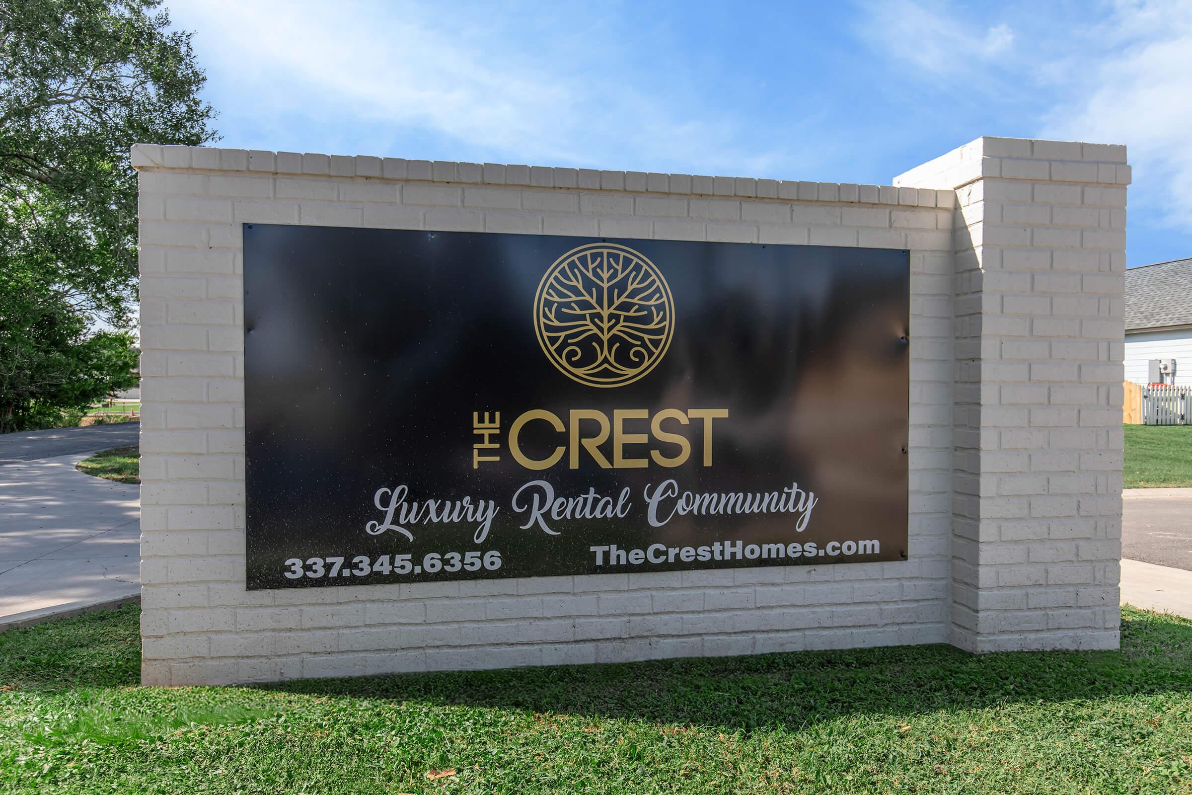 Sign for The Crest Luxury Rental Community, featuring the logo of a tree, with contact information and website link. The sign is mounted on a brick structure and is situated in a green grassy area.