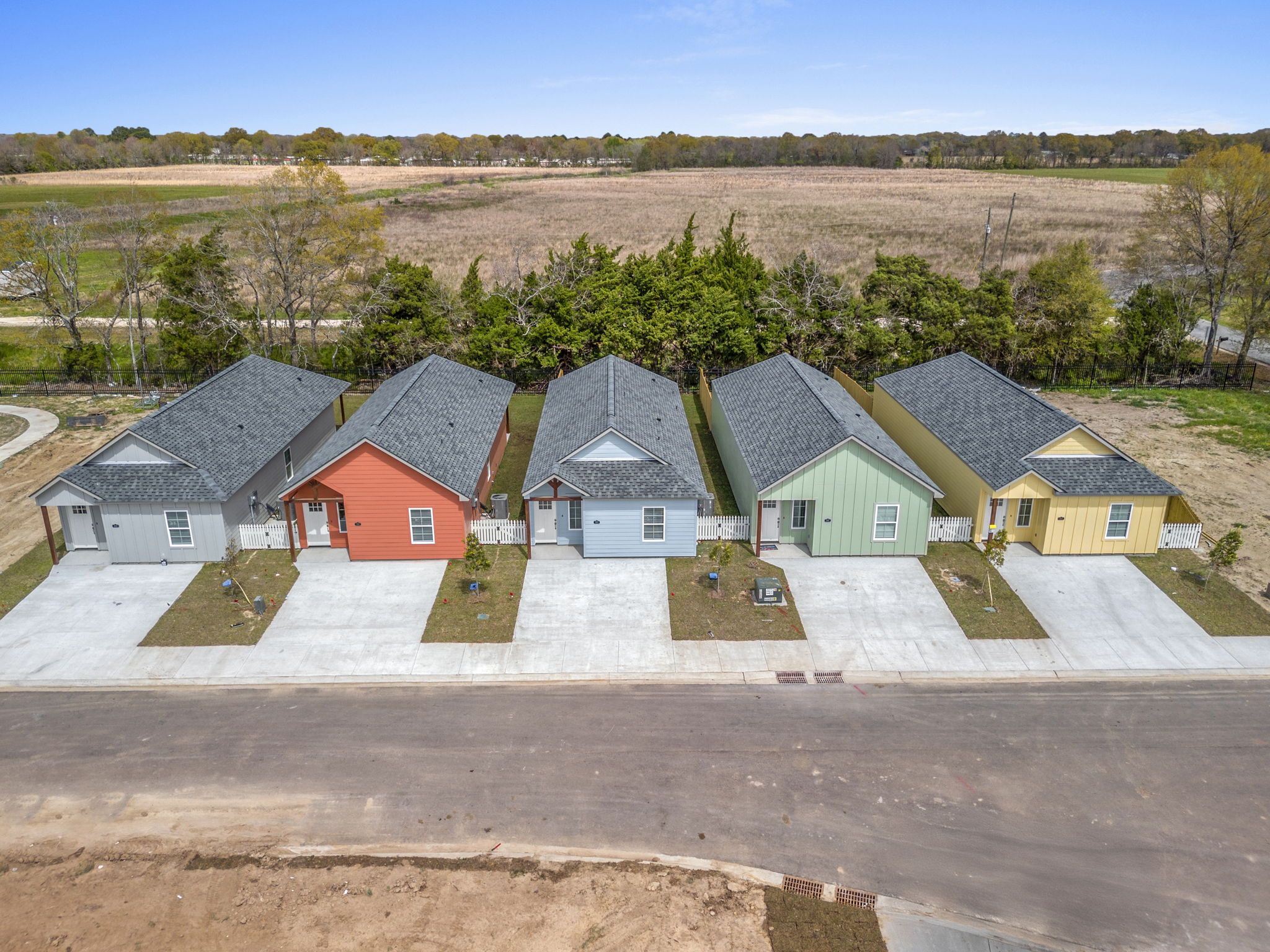 A row of modern single-family homes in various colors, including red, gray, green, and yellow, situated on a paved street. Each house features a front yard with small decorations and landscaping. The background includes trees and an open field under a clear blue sky.