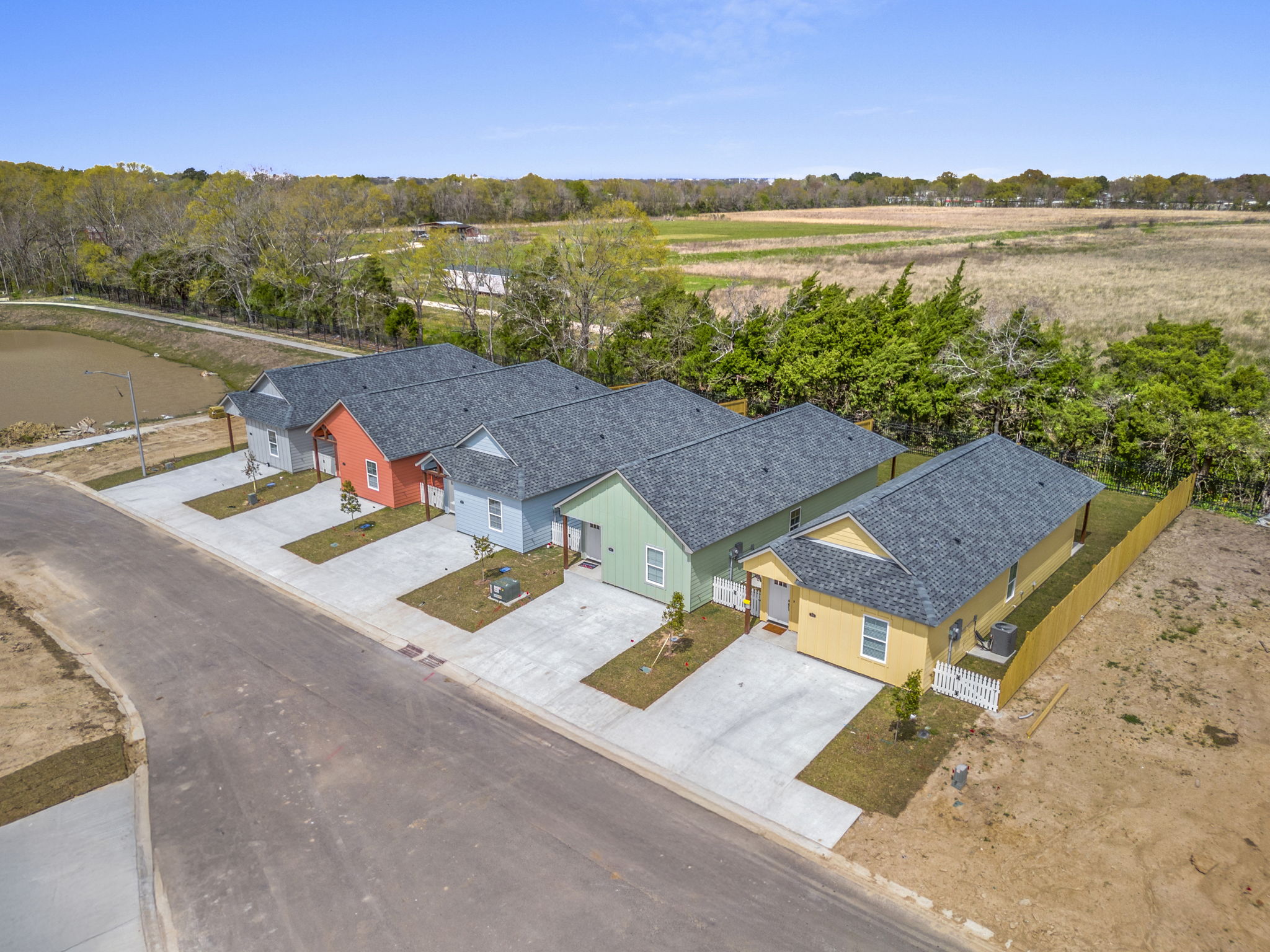 Aerial view of a row of colorful single-story homes on a paved street, with green grass and trees in the background. The houses feature different colors: red, blue, green, and yellow. A grassy area extends behind the homes, and the scene is clear and bright under a blue sky.