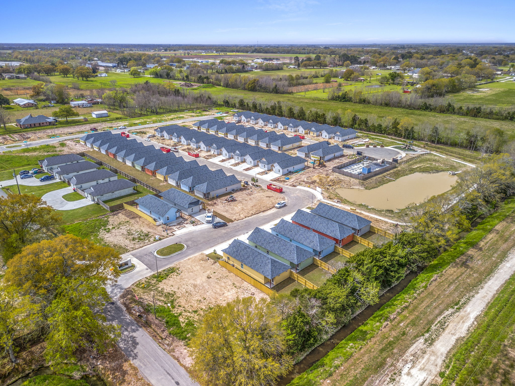 Aerial view of a new residential development featuring several rows of single-family homes, a central pond, and green spaces. The area is surrounded by fields and trees, showcasing a blend of urban living and nature. Some homes are under construction, and vehicles are parked along the streets.