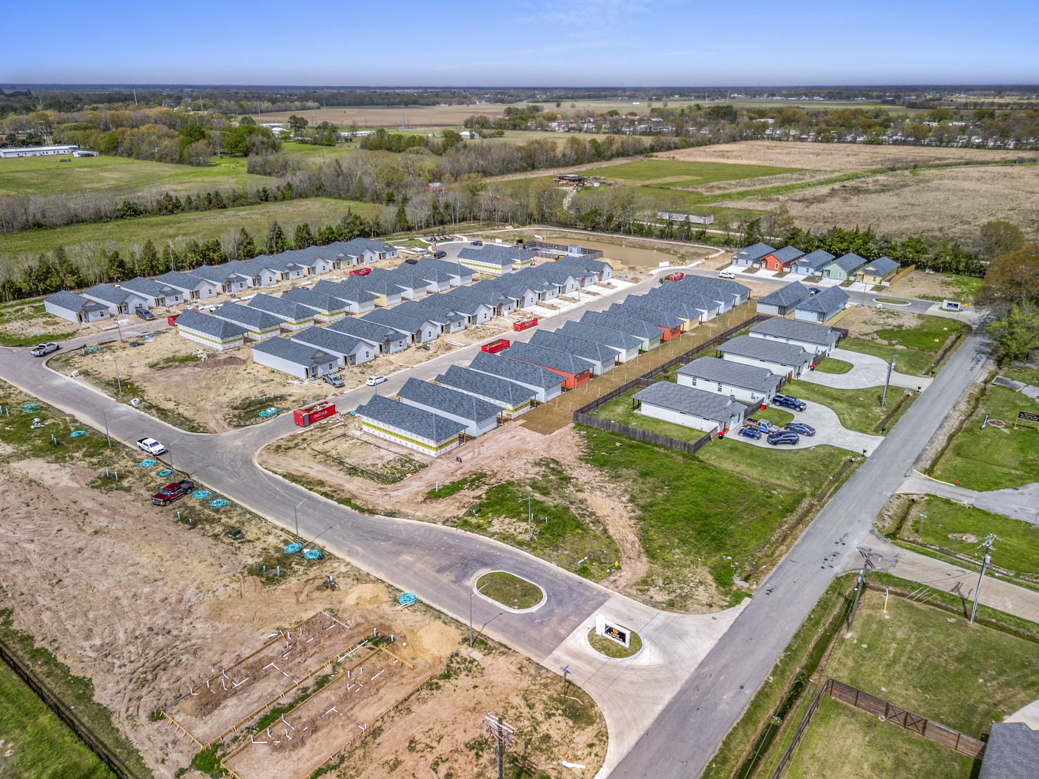 Aerial view of a residential development featuring a series of newly constructed single-family homes arranged in rows. The surrounding area includes open fields and a few trees, with roads linking the houses. Some plots appear unfinished or under construction, showcasing a mix of landscaping and bare earth.