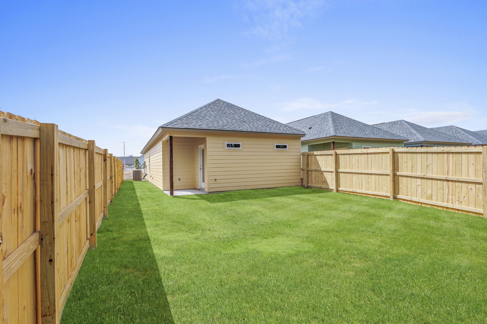A well-maintained backyard featuring a neatly mowed green lawn surrounded by wooden fences. In the background, a single-story house with a gray roof and windows is visible. The sky above is clear with a few clouds, creating a bright and sunny atmosphere.