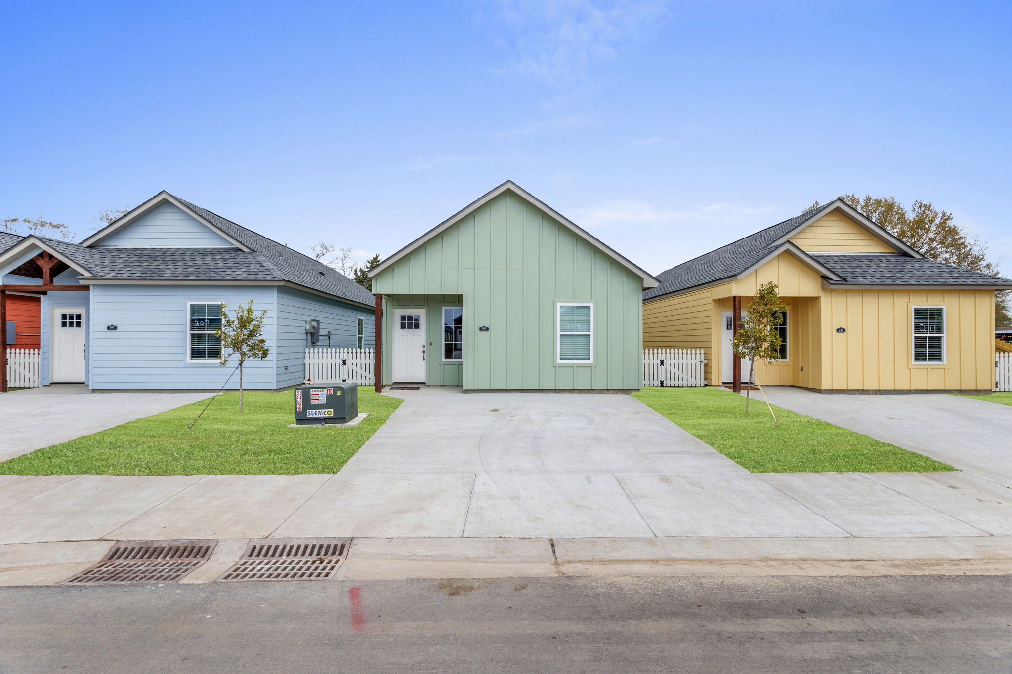 Three brightly colored houses lined up side by side on a grassy lawn. The houses feature a blue house on the left, a green house in the middle, and a yellow house on the right. Each house has a front door, windows, and is set against a clear blue sky. A for sale sign is visible in front of the green house.