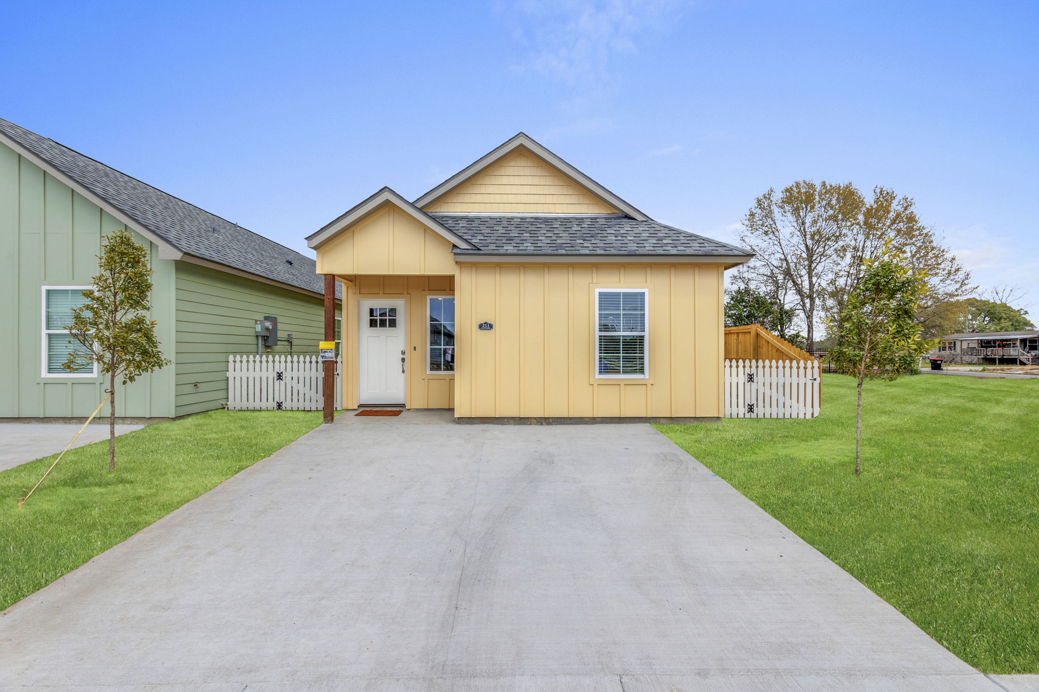 A charming single-story house with a yellow exterior, a front porch, and neatly landscaped grass. The house features a gabled roof and large windows. Flanking the home are green painted houses, and there are small trees planted in the front yard, creating a welcoming atmosphere.