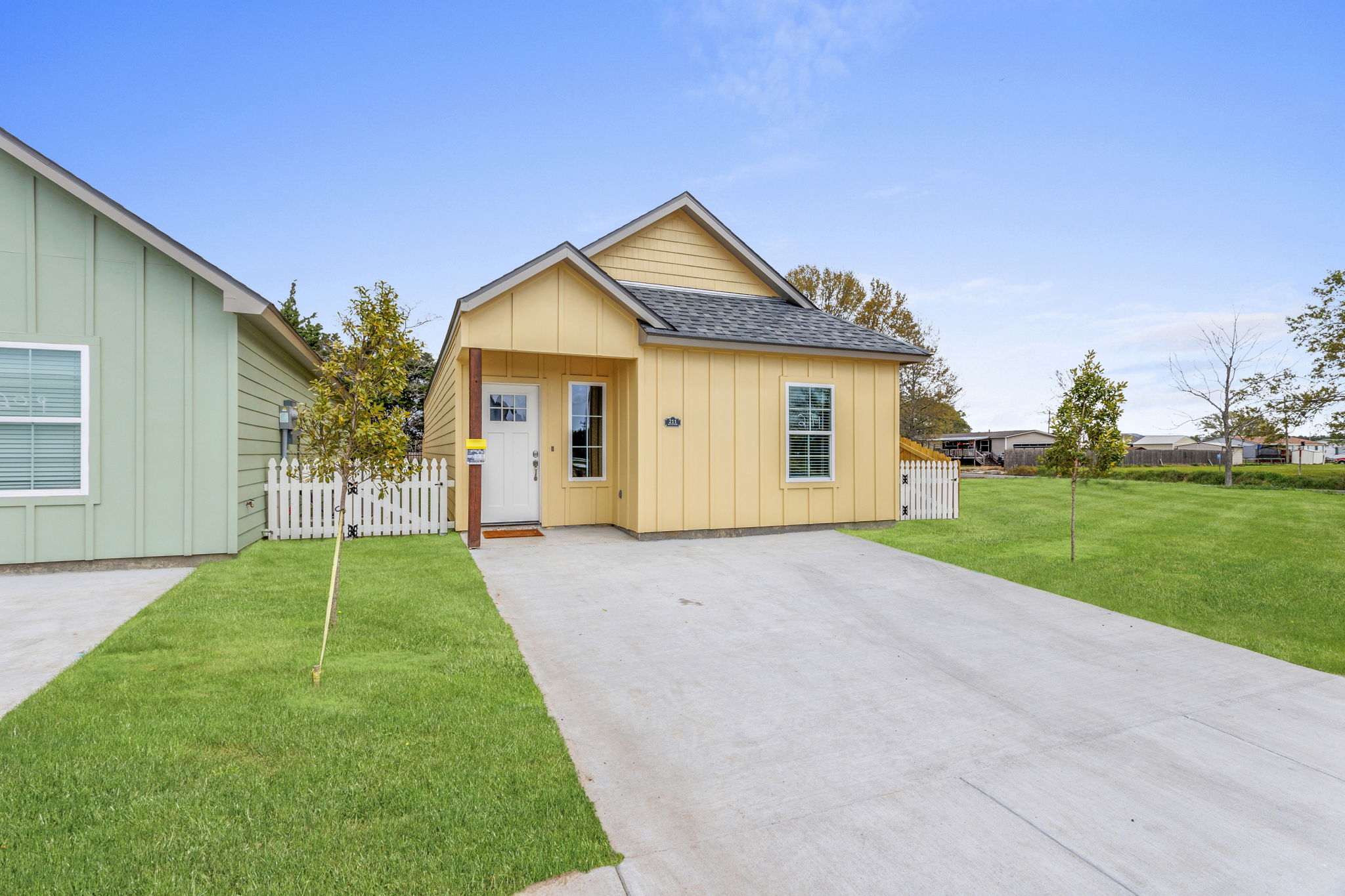 A small, single-story house with a yellow exterior and gray roof sits on a driveway. The house features a front door with a welcoming porch, surrounded by a well-maintained lawn. To the left, there is a green house, and in the background, additional houses and trees are visible under a clear blue sky.