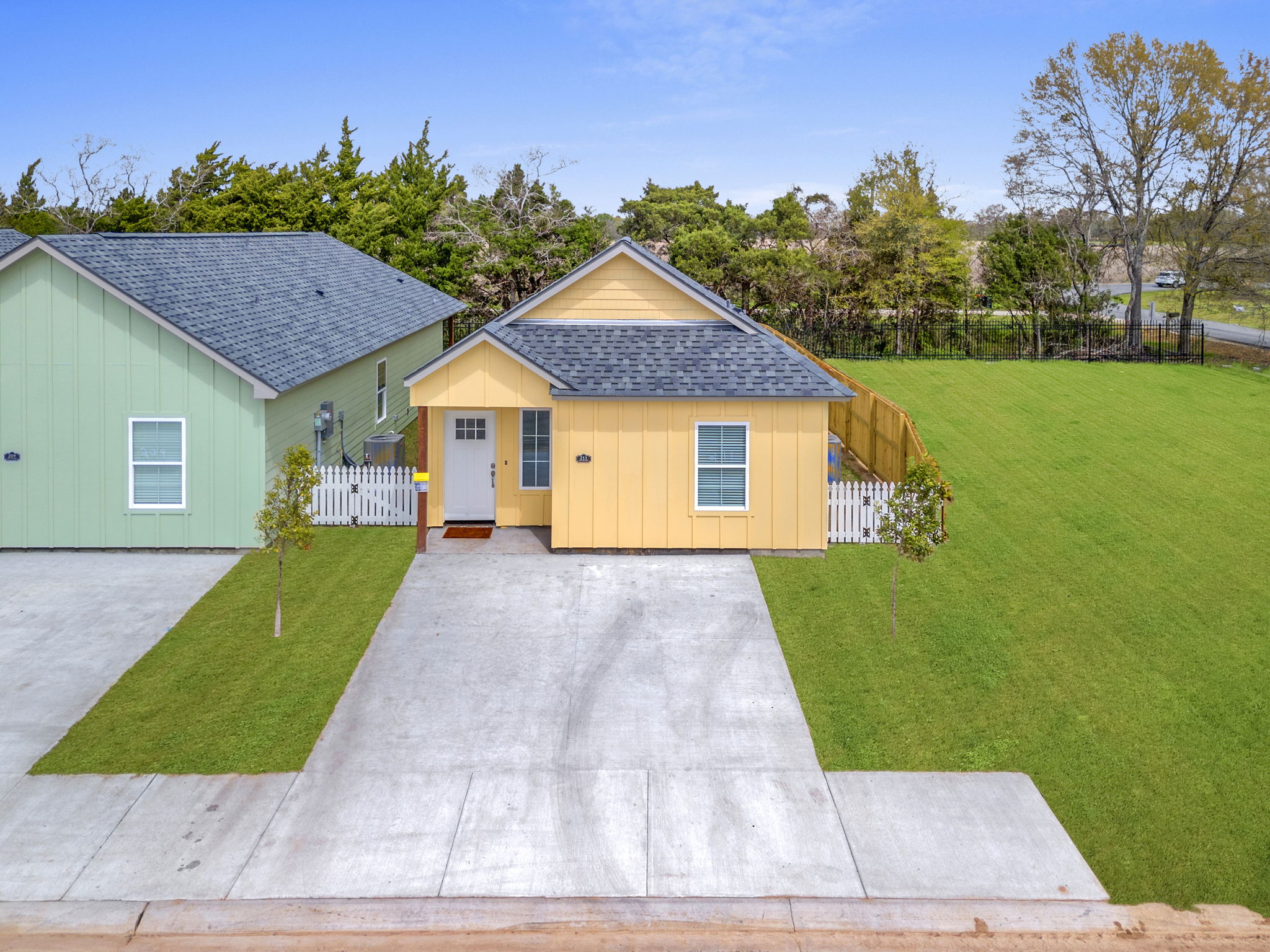 A yellow house with a gray roof and white trim, situated between a green house and a grassy area. The front yard features a small fence and two young trees. The driveway is paved and leads up to the entrance of the house, surrounded by lush greenery.